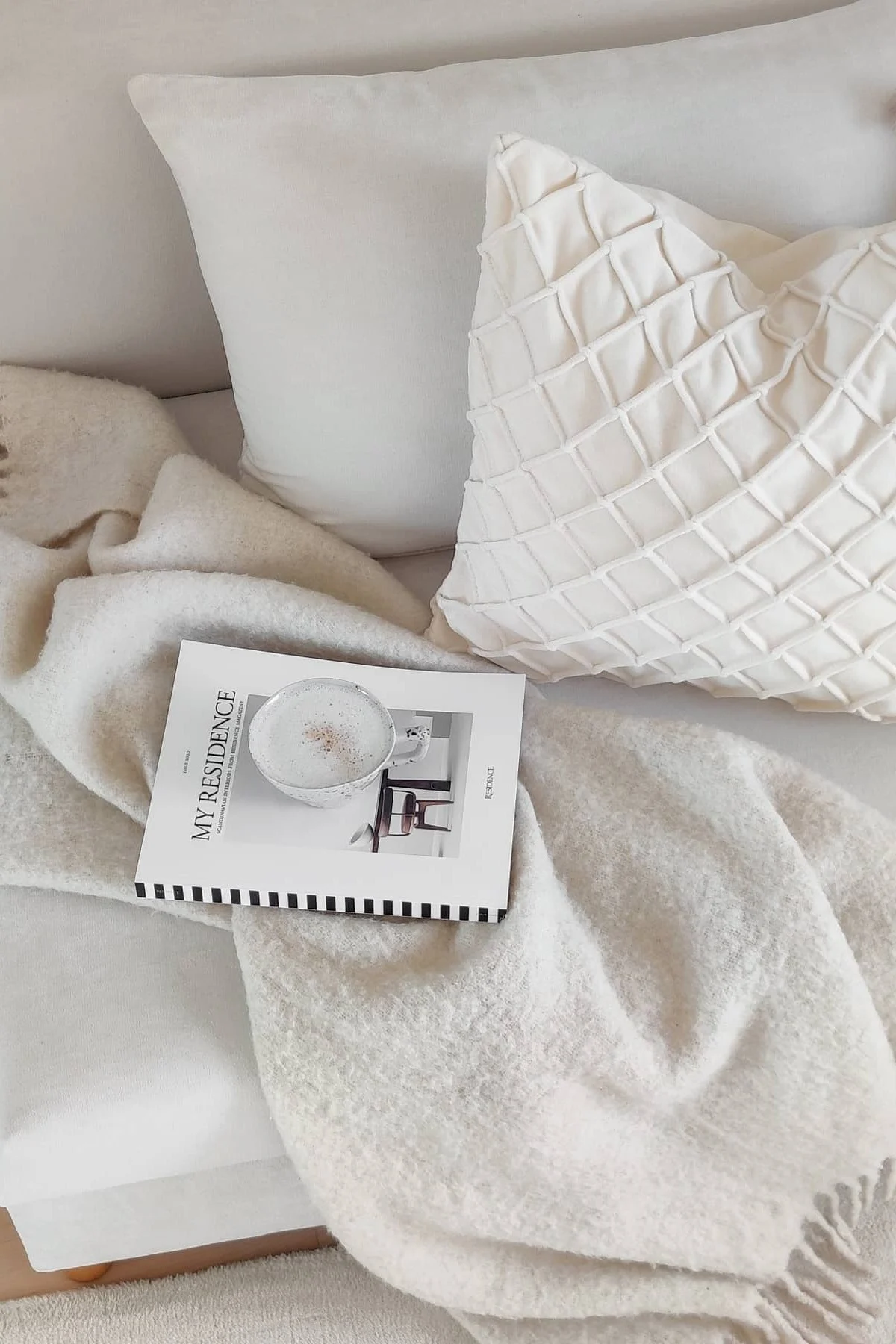 Regalos de San Valentín: sobre un sofá blanco hay dos cojines blancos de Chhatwal & Jonsson, la manta Ralph de Klippan Yllefabrik, un libro y una taza de café.