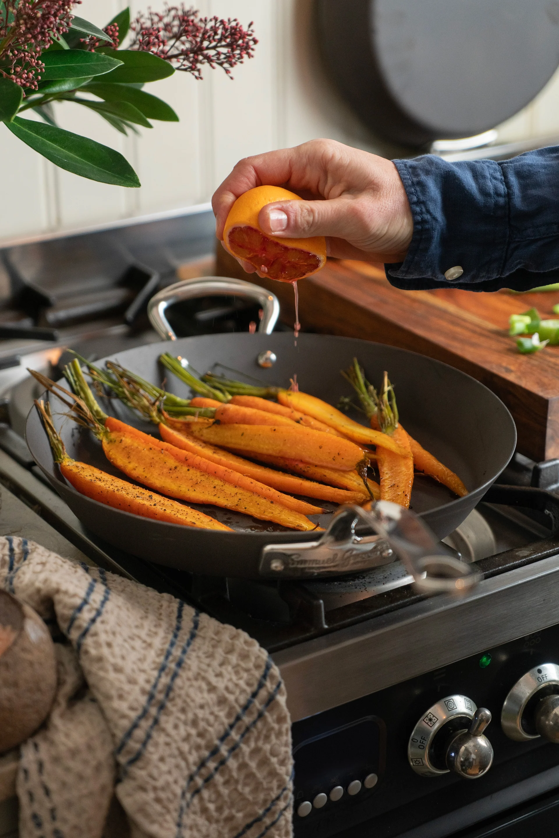 Una sartén de acero al carbono con zanahorias está sobre la cocina, mientras una mano exprime el jugo de un cítrico sobre ellas.