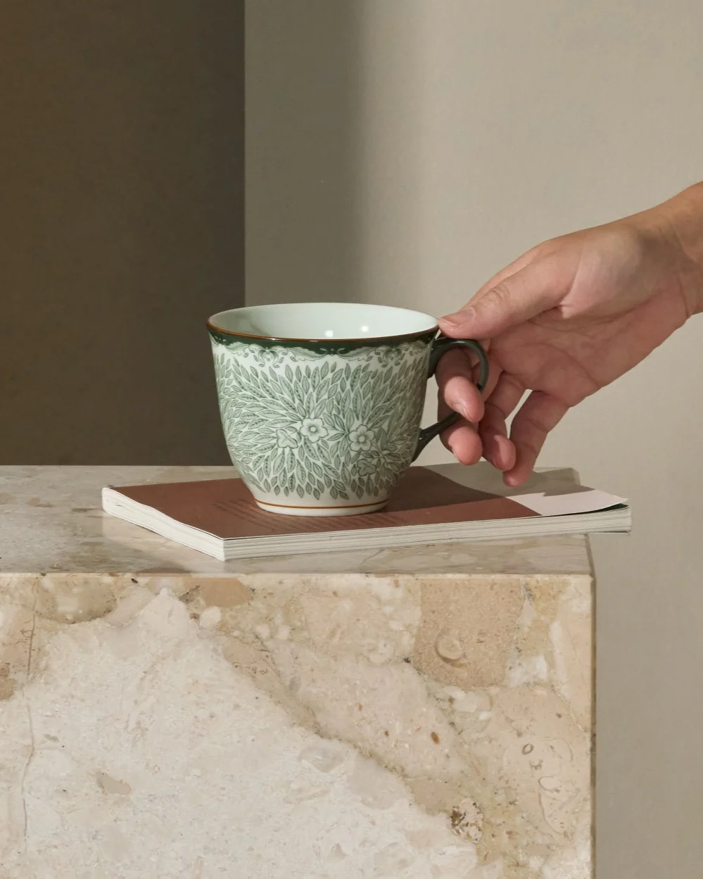 Persona alcanzando una taza verde estampada en una mesa auxiliar de mármol junto a un sofá.