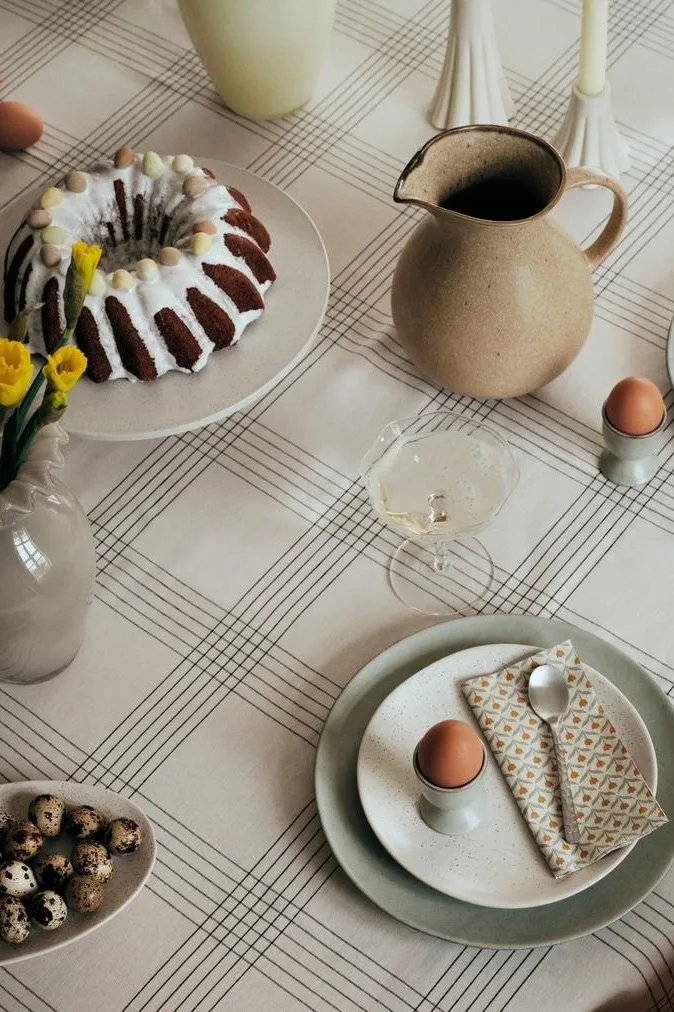 Mesa puesta para Pascua con un bizcocho, narcisos, huevos y bebidas sobre un mantel a cuadros.