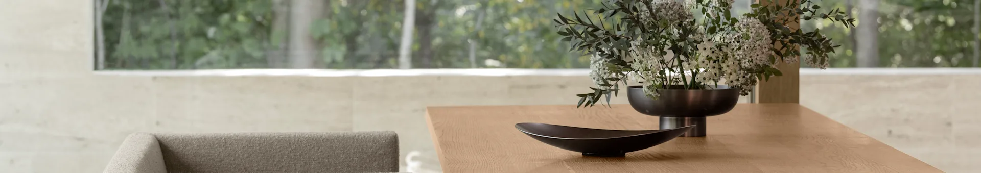 Mesa de comedor de madera moderna con un cuenco decorativo oscuro y un jarrón con flores blancas y verdes, con una vista exterior borrosa a través de una gran ventana.