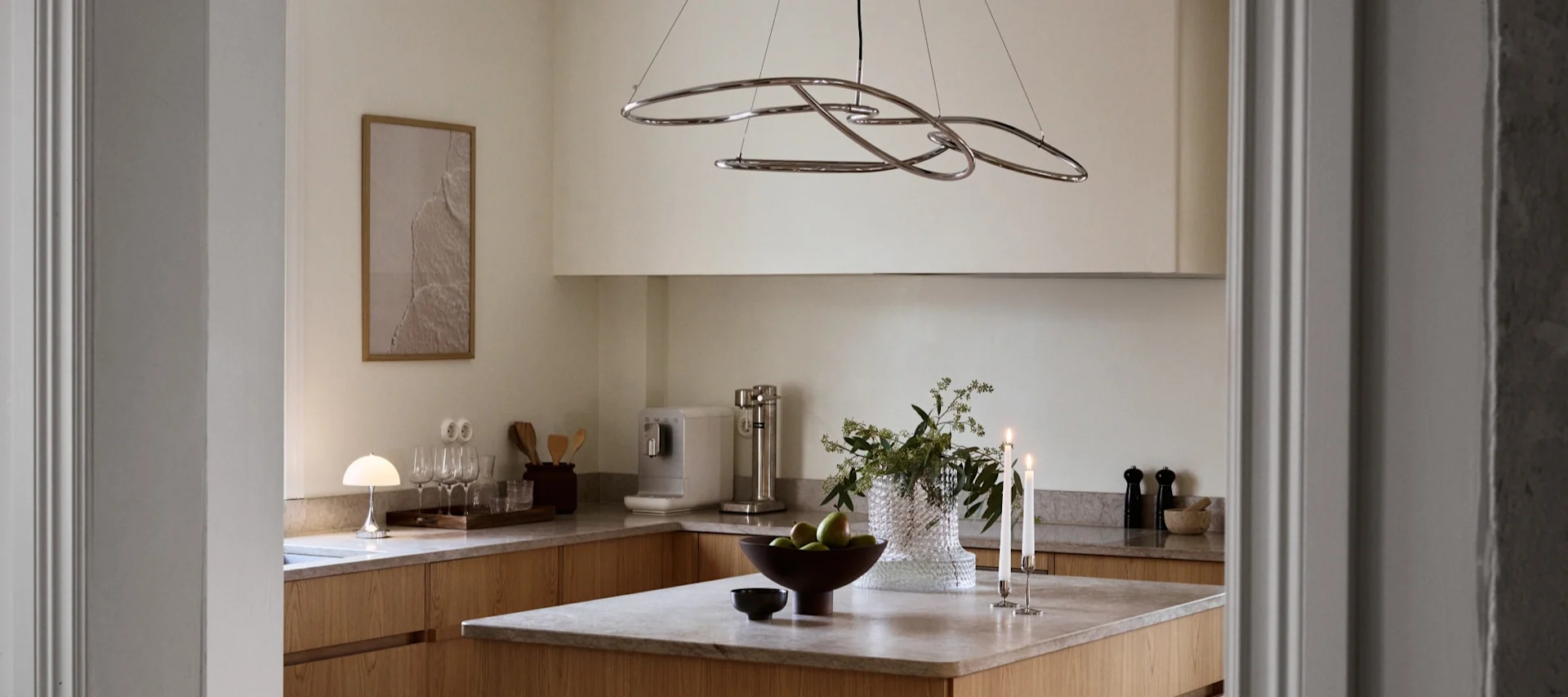 Cocina moderna con muebles de madera clara, isla con manzanas verdes, velas encendidas y lámpara escultural de cromo.