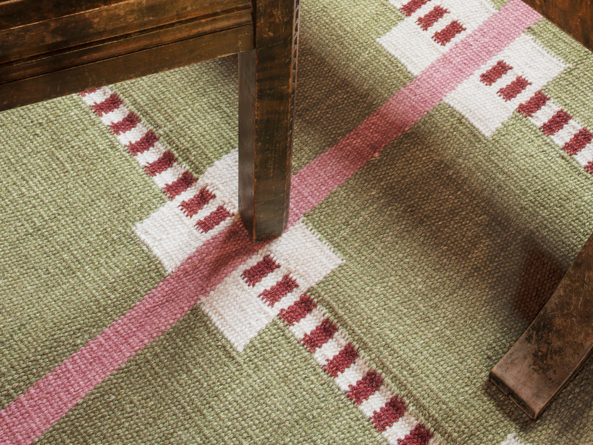 Una alfombra con patrón geométrico verde, blanco y rojo, y una pata de mueble de madera oscura proyectando una sombra.