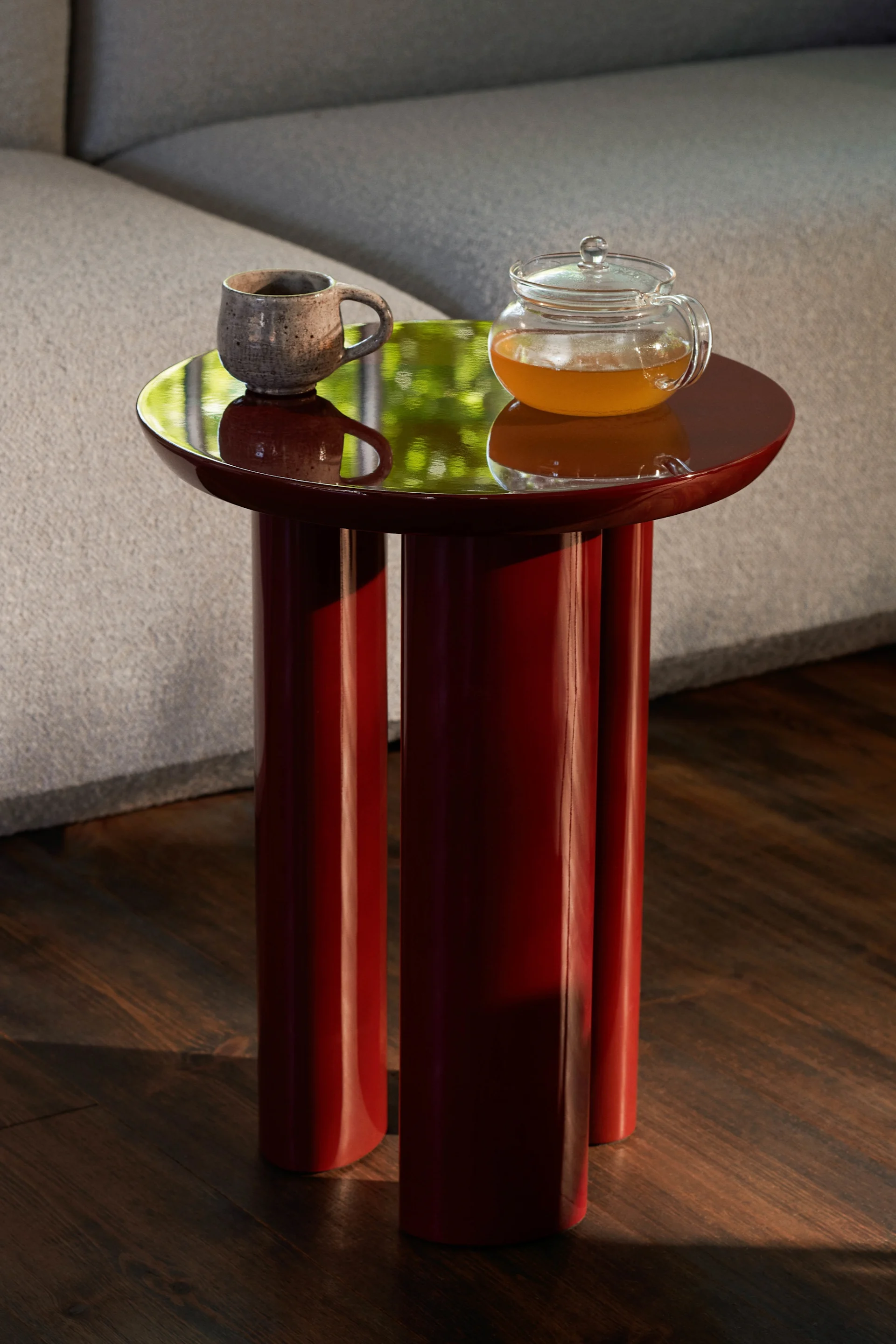 Mesa auxiliar roja brillante con taza y tetera. Reflejos verdes en la superficie, junto a sofá gris y suelo de madera.