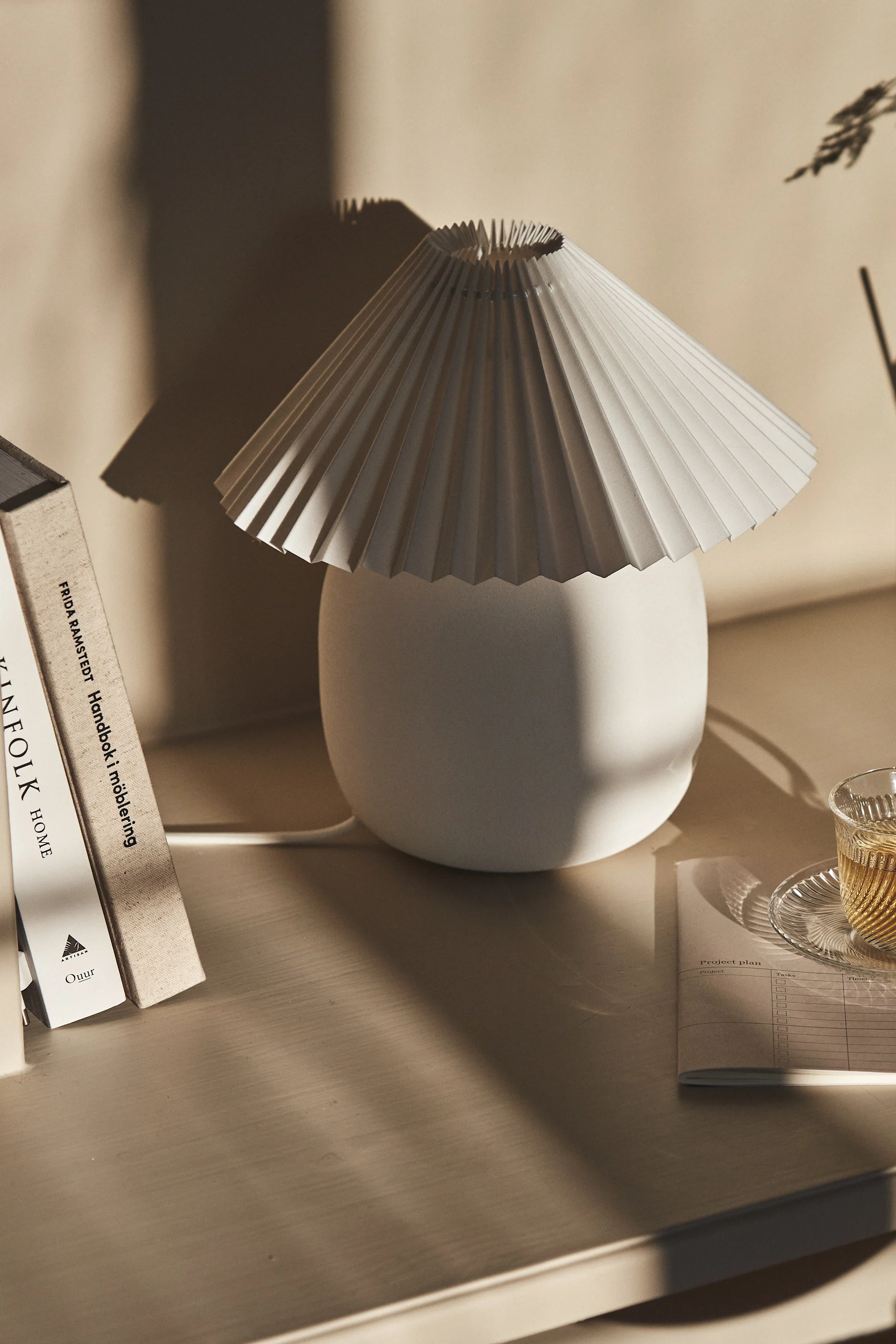 Una lámpara de mesa blanca plisada junto a libros y una taza de té en un escritorio de madera clara, con sombras suaves.