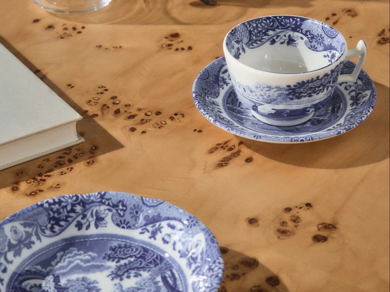 Taza y platillo de té con patrón azul y blanco junto a un plato a juego sobre una mesa de madera clara con nudos, con un libro blanco visible al fondo.