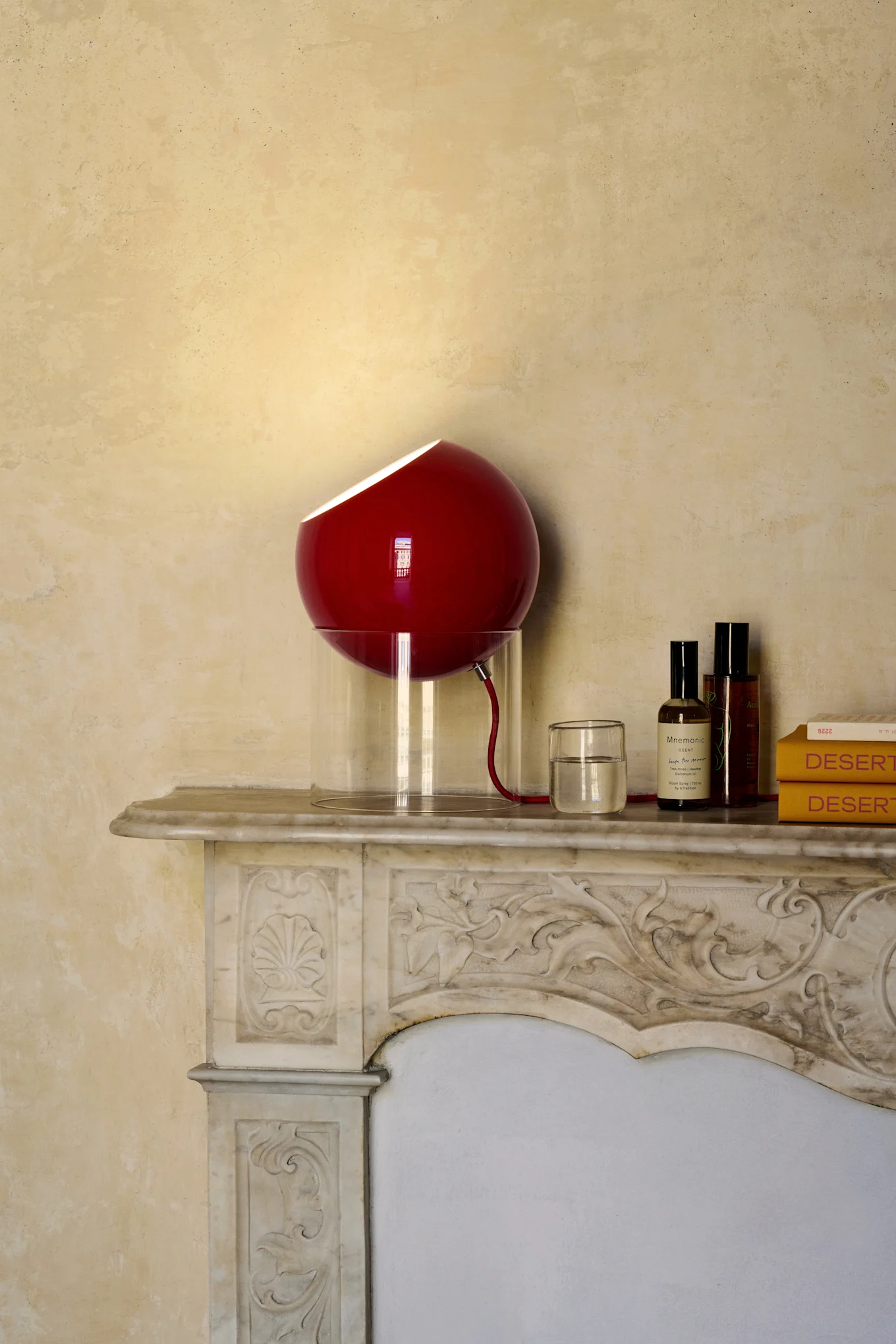 Lámpara esférica roja con base translúcida sobre chimenea de mármol decorada, junto a un vaso de agua, dos botellas y dos libros amarillos.