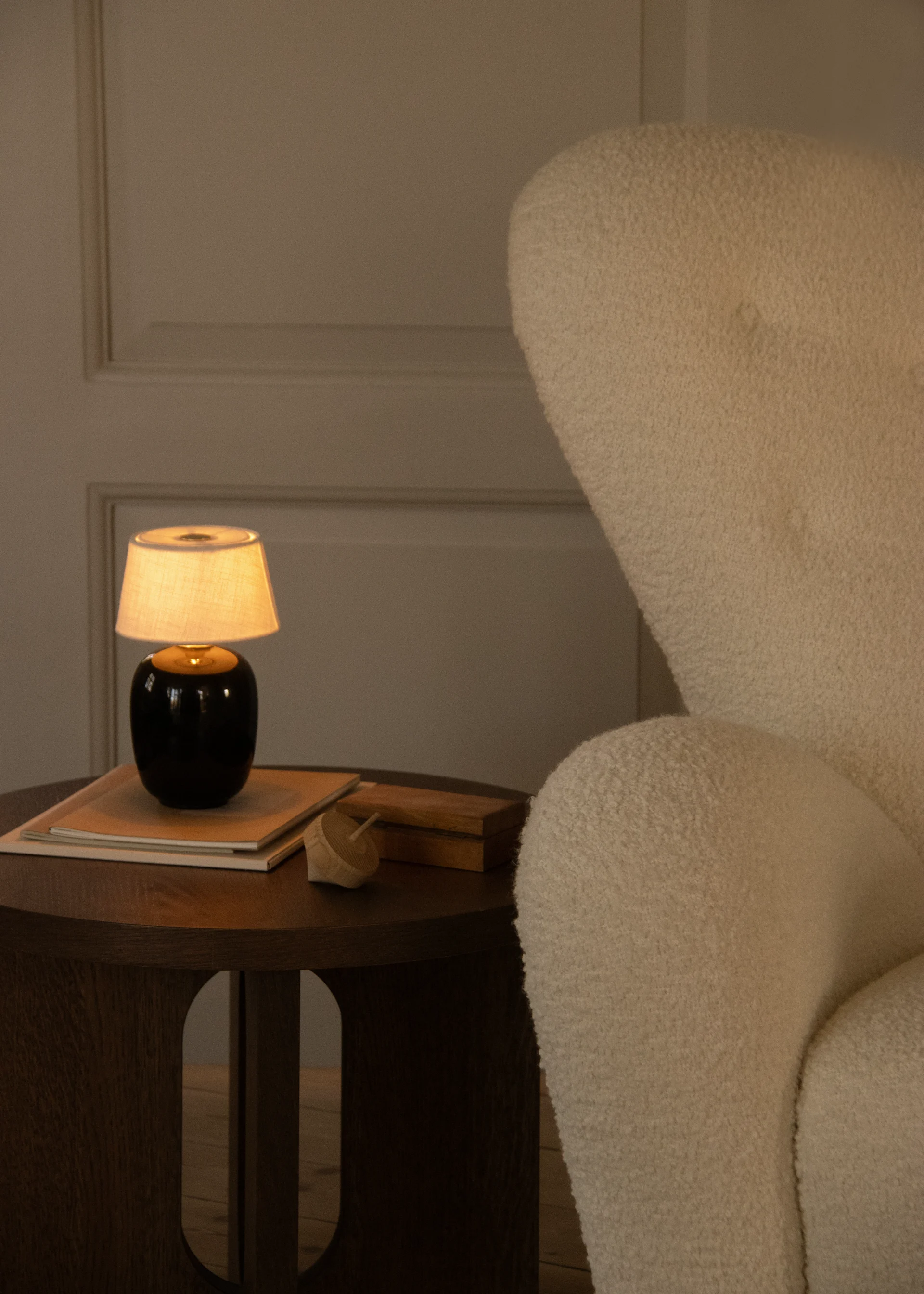 Acogedor rincón cálidamente iluminado con un sillón texturizado crema y una mesa auxiliar de madera con una lámpara, libros y un juguete.