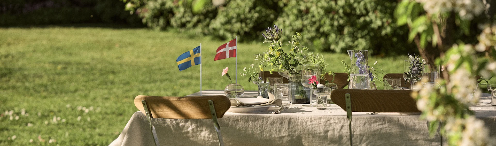 Mesa de exterior con banderas sueca y danesa, flores silvestres y cristalería en un jardín soleado.