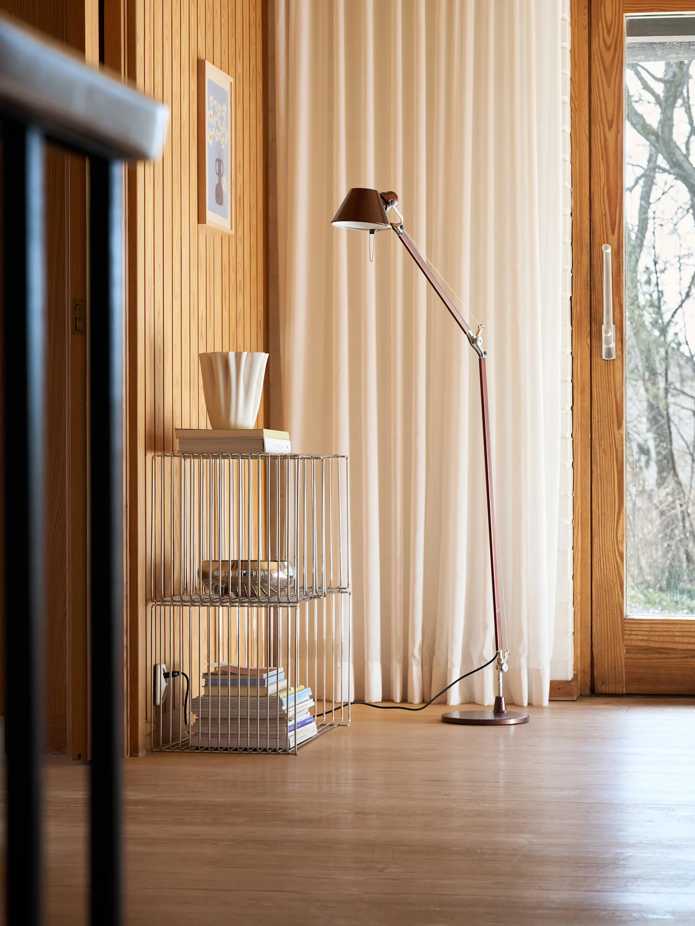 Una acogedora habitación con paredes y suelo de madera, una lámpara de pie roja, una estantería cromada con libros y un jarrón blanco estriado, y una ventana con cortinas claras.
