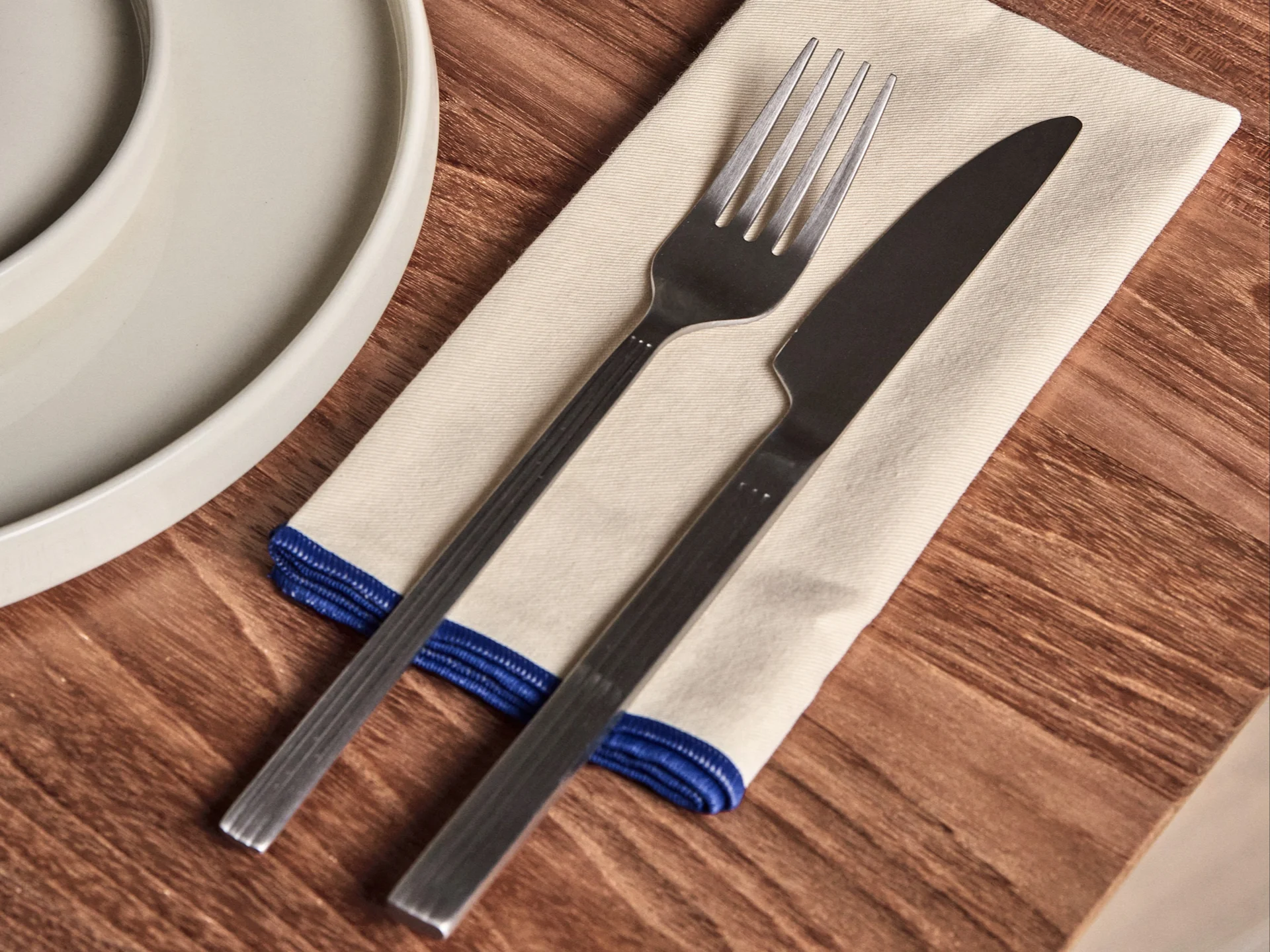 Tenedor y cuchillo sobre una servilleta color crema con borde azul, junto a un plato blanco en una mesa de madera.