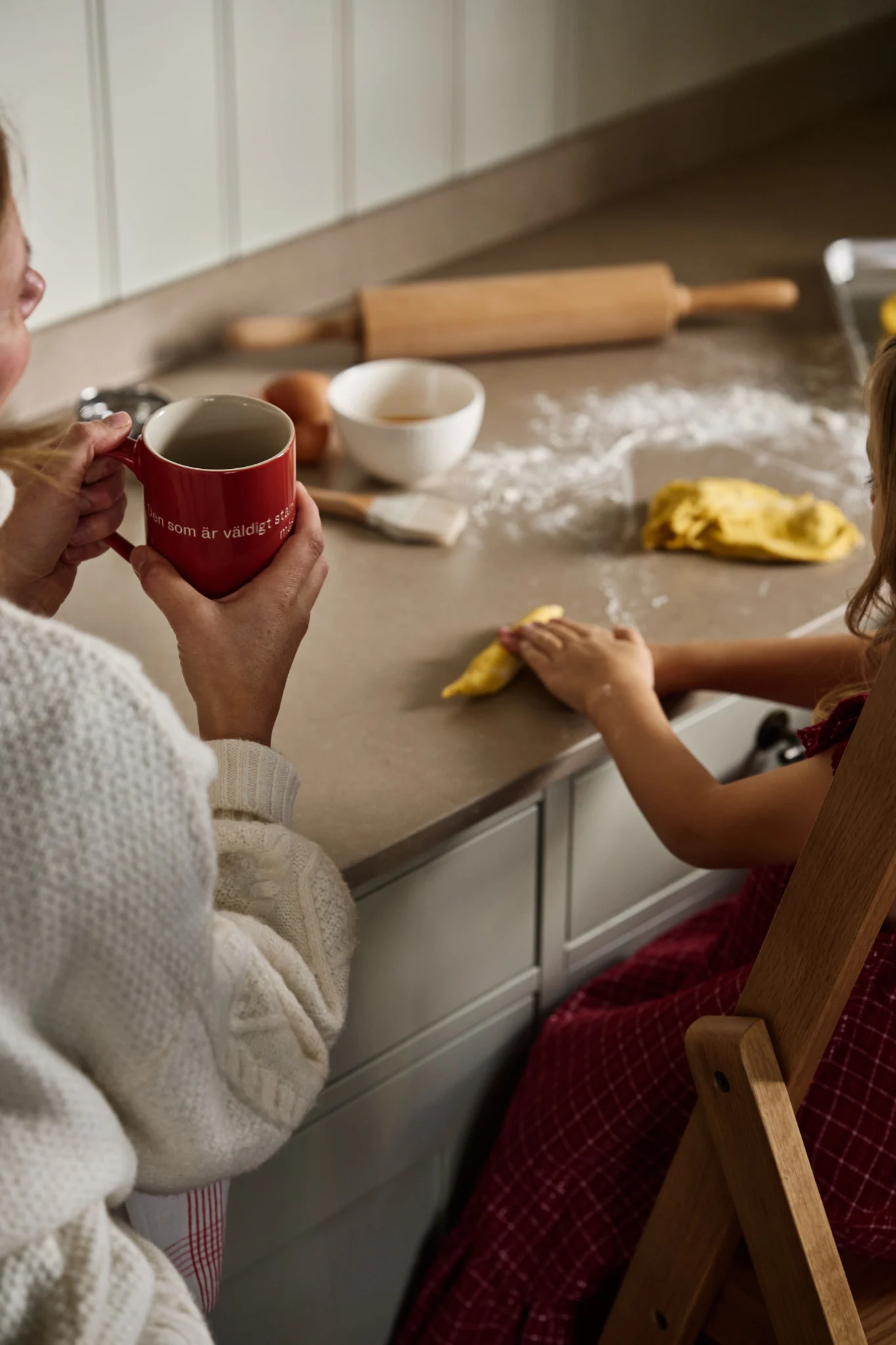 Una mujer está sentada en la encimera con una taza de Astrid Lindgren, frente a ella una niña que amasa la masa.