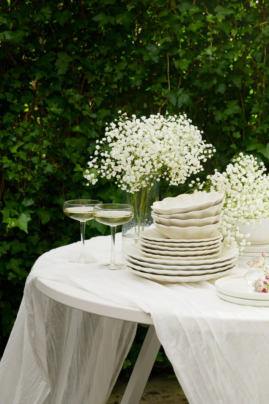 Mesa exterior puesta con vajilla blanca, copas de champán y flores de paniculata sobre follaje verde.