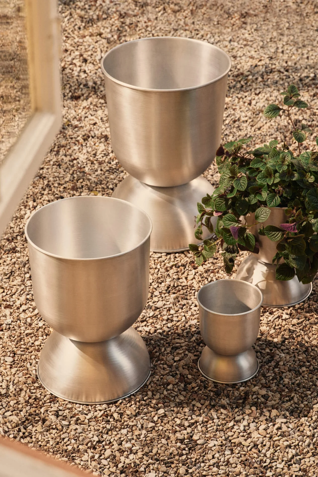 Tres jardineras de metal cepillado de diferentes tamaños sobre guijarros, una con una planta verde.