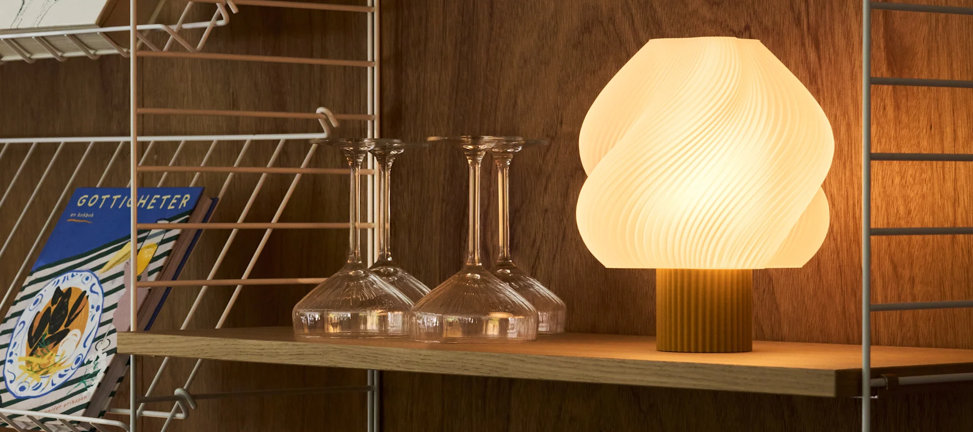 Una lámpara ovalada acanalada e iluminada y varios vasos transparentes se encuentran en un carrito de bar de madera frente a una pared cálida y texturizada.