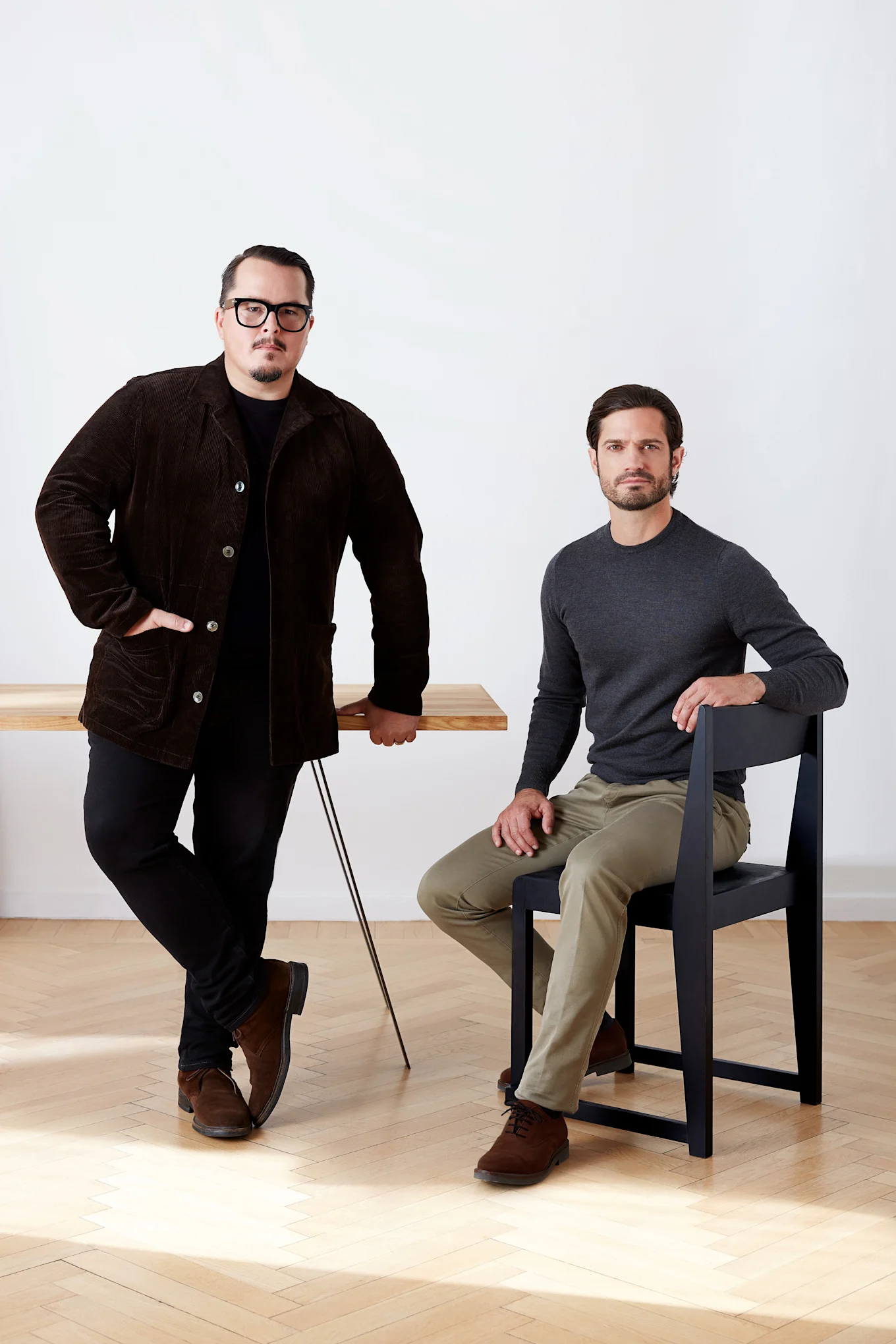 Dos hombres, uno de pie junto a una mesa de madera con una chaqueta de pana marrón, el otro sentado en una silla negra con una camisa gris.