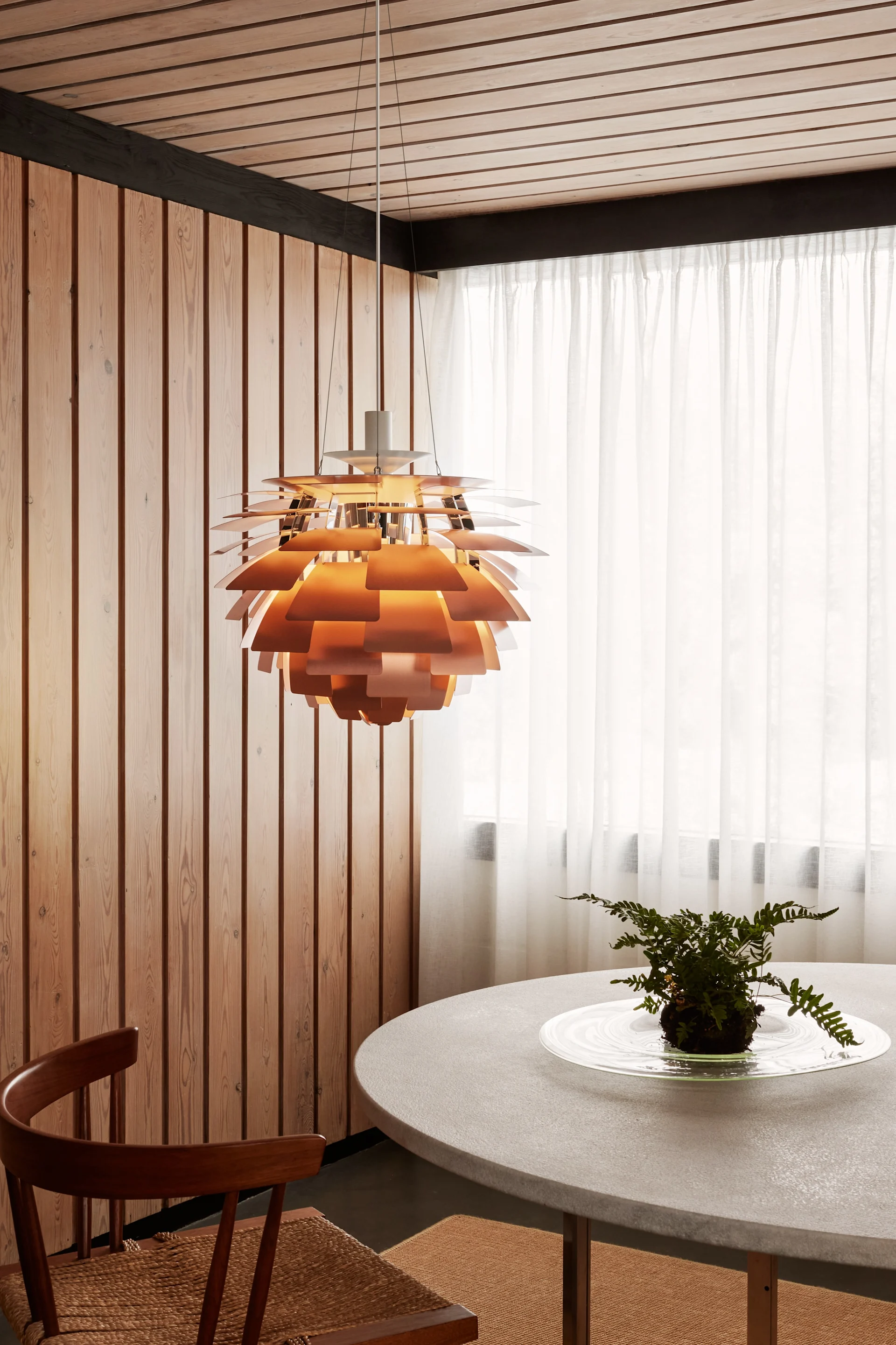 Una lámpara colgante de cobre en capas cuelga sobre una mesa redonda gris con un helecho, junto a paredes de madera y cortinas blancas transparentes.