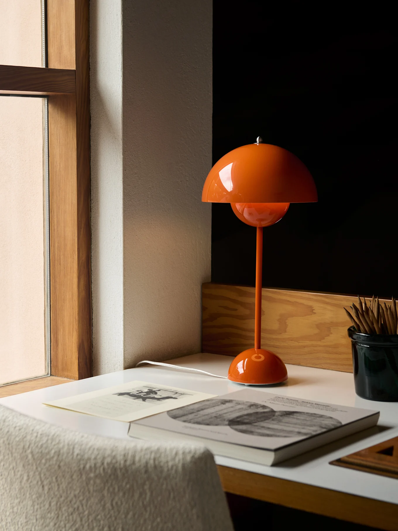 Una lámpara de mesa naranja con forma de seta sobre un escritorio blanco junto a una ventana, con libros y lápices.