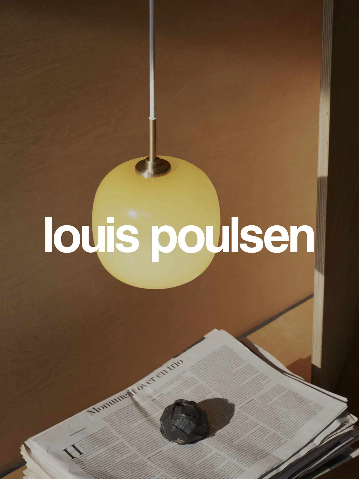 Logotipo de Louis Poulsen superpuesto en una escena cálida con una lámpara colgante esférica amarilla, periódicos apilados y un objeto decorativo oscuro.