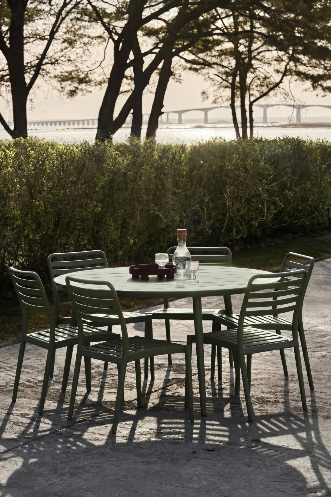 Conjunto de comedor verde al aire libre con mesa, cinco sillas, botella de agua y vasos en patio. Puente lejano.