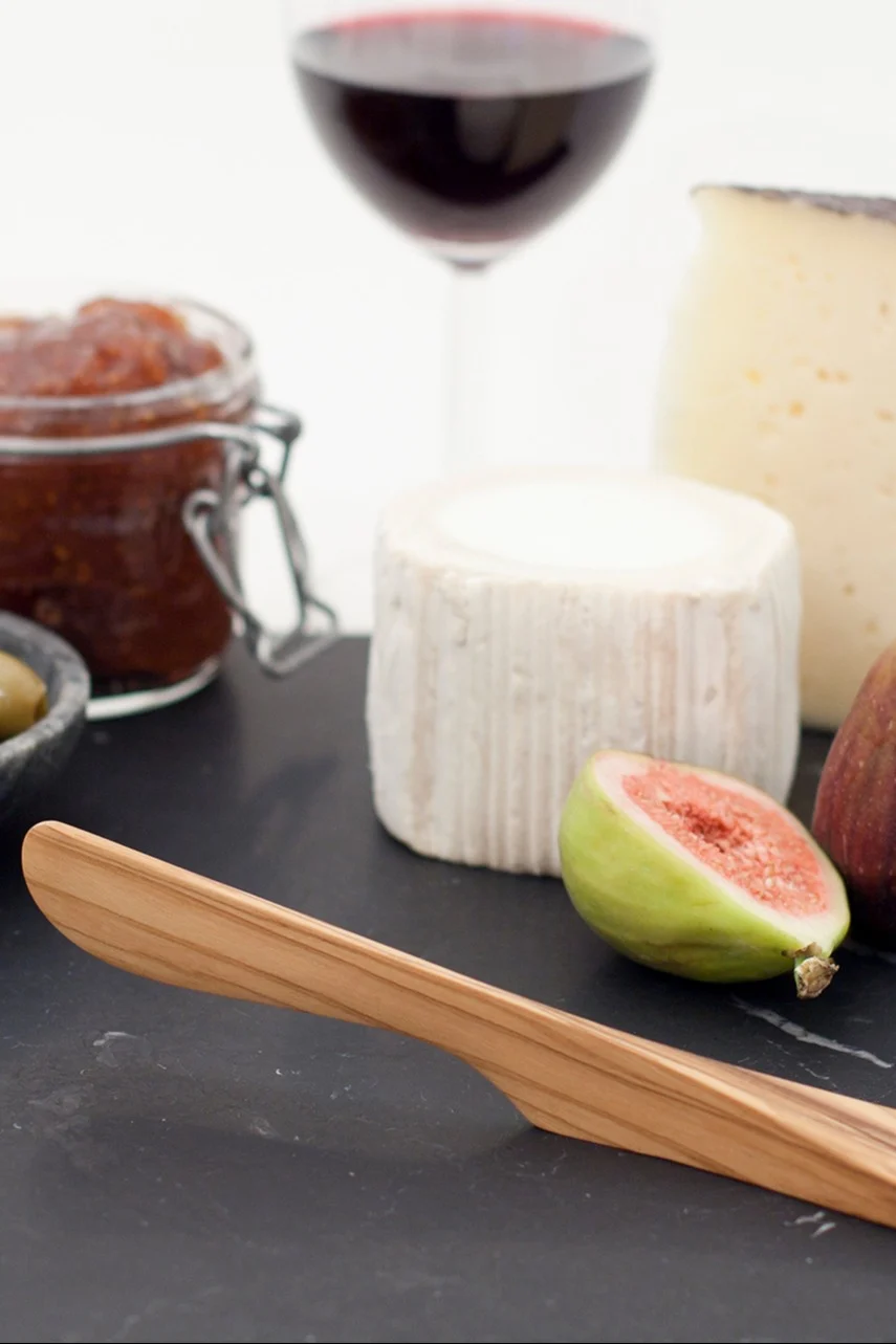 Una tabla de quesos con un cuchillo de madera, dos quesos, un higo partido por la mitad, mermelada, aceitunas y una copa de vino tinto.