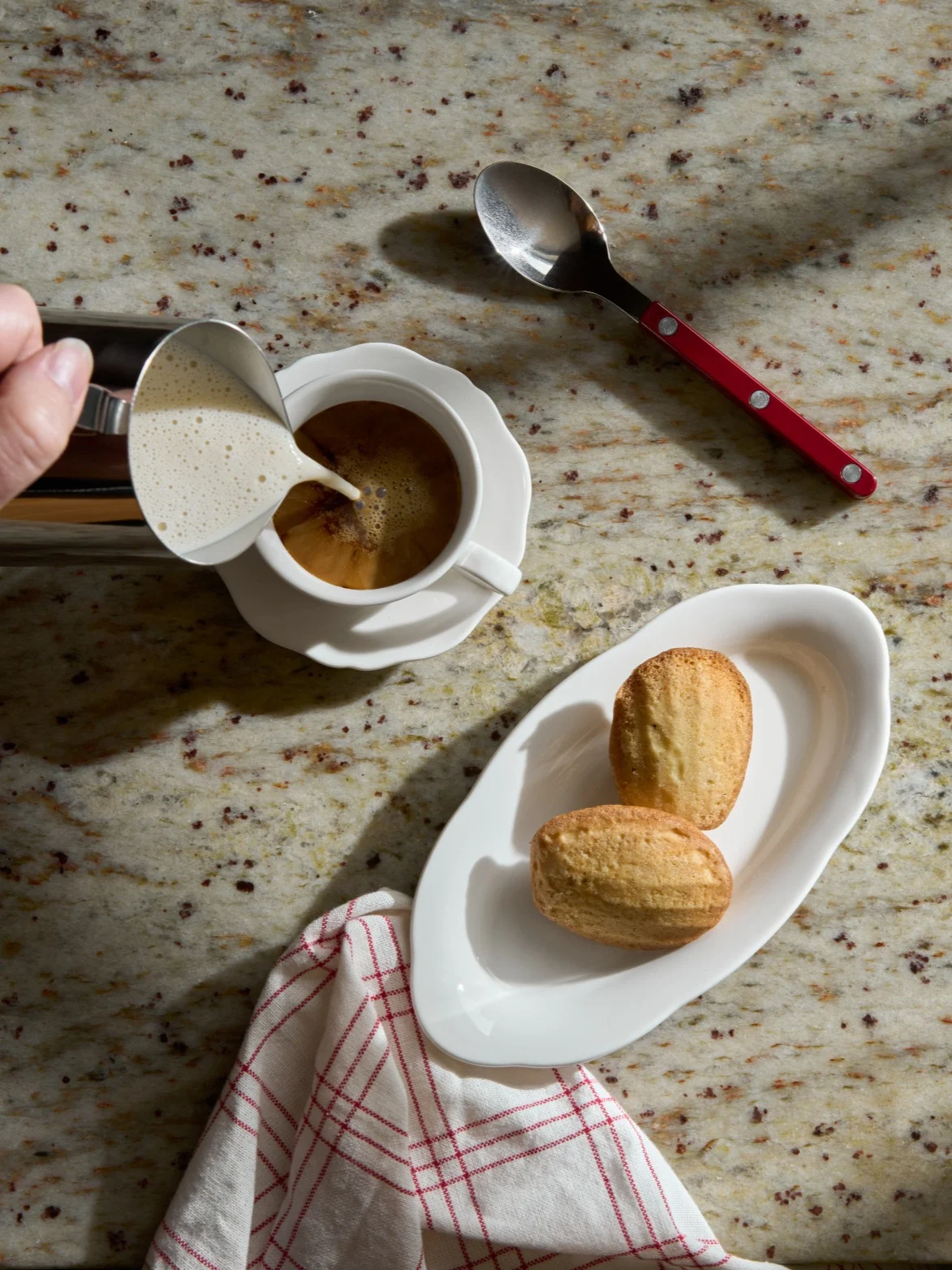 Mano vertiendo leche espumada en una taza de café junto a magdalenas en una encimera de granito.