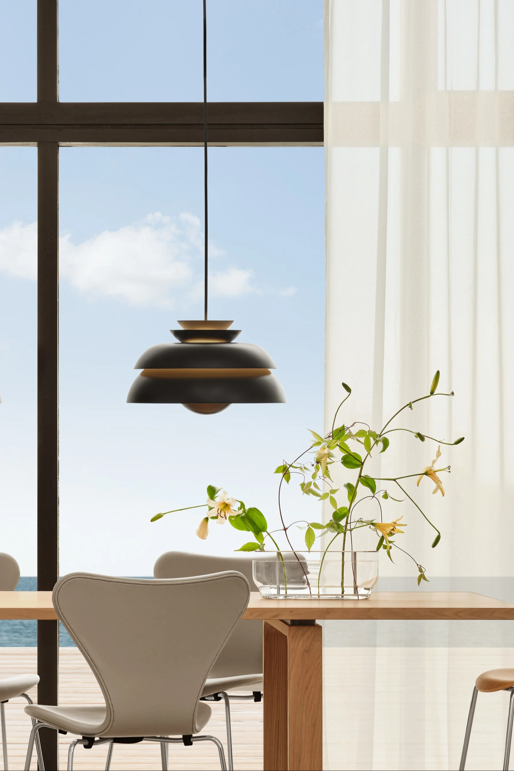 Comedor: mesa de madera, sillas, lámparas oscuras. Flores. Ventana, cortinas transparentes, vista al mar.