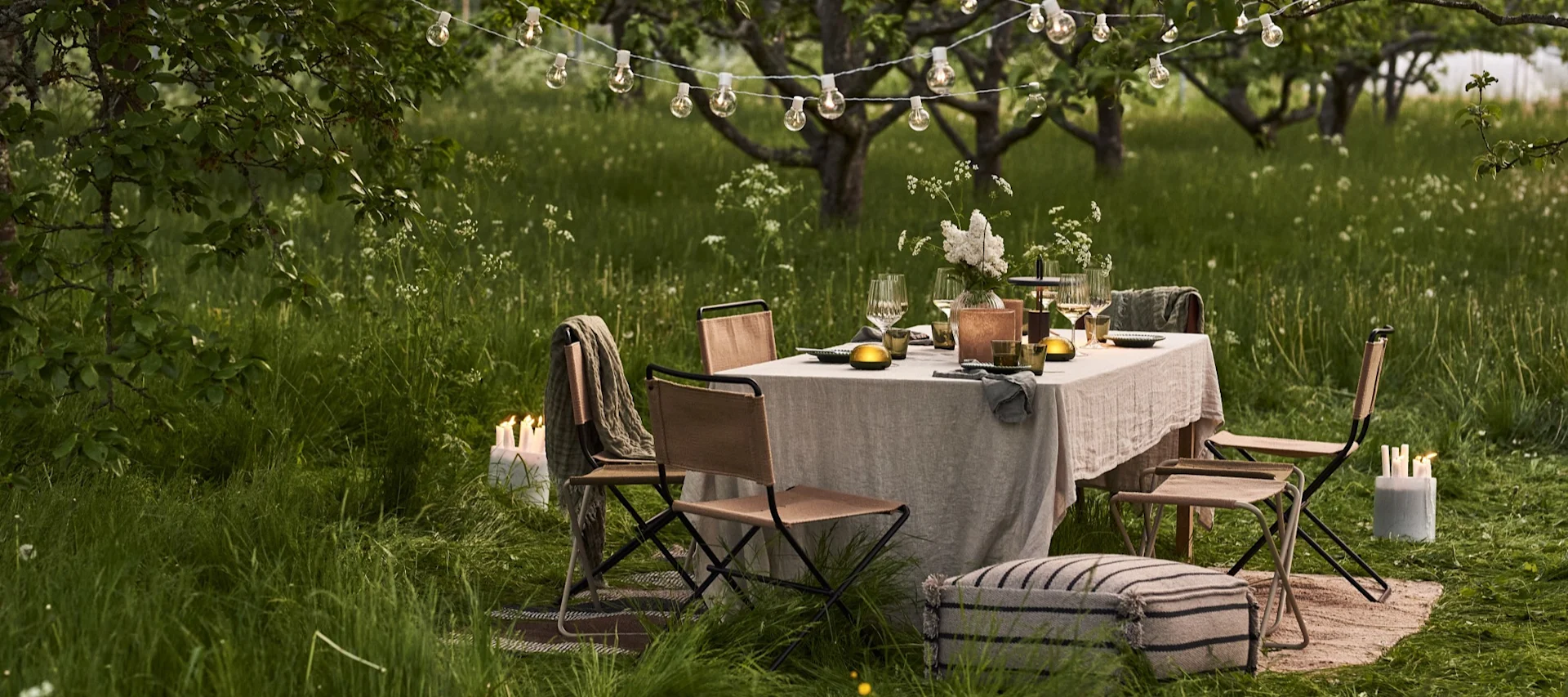 Mesa de comedor al aire libre bajo luces de cuerda en un jardín con hierba al anochecer, con sillas y velas.