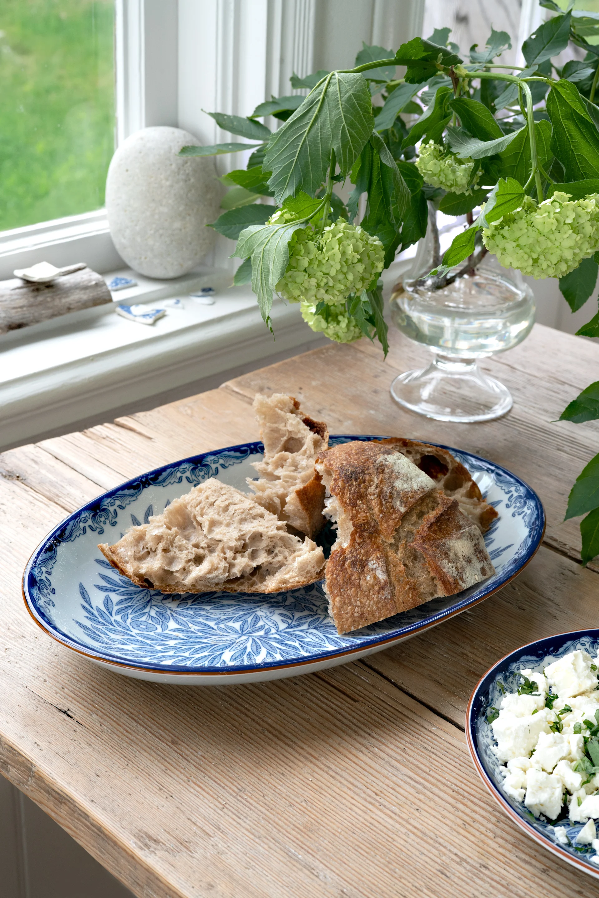 Trozos de pan rústico rasgados en un plato azul y blanco con estampado, con queso desmenuzado y hortensias verdes sobre una mesa de madera.