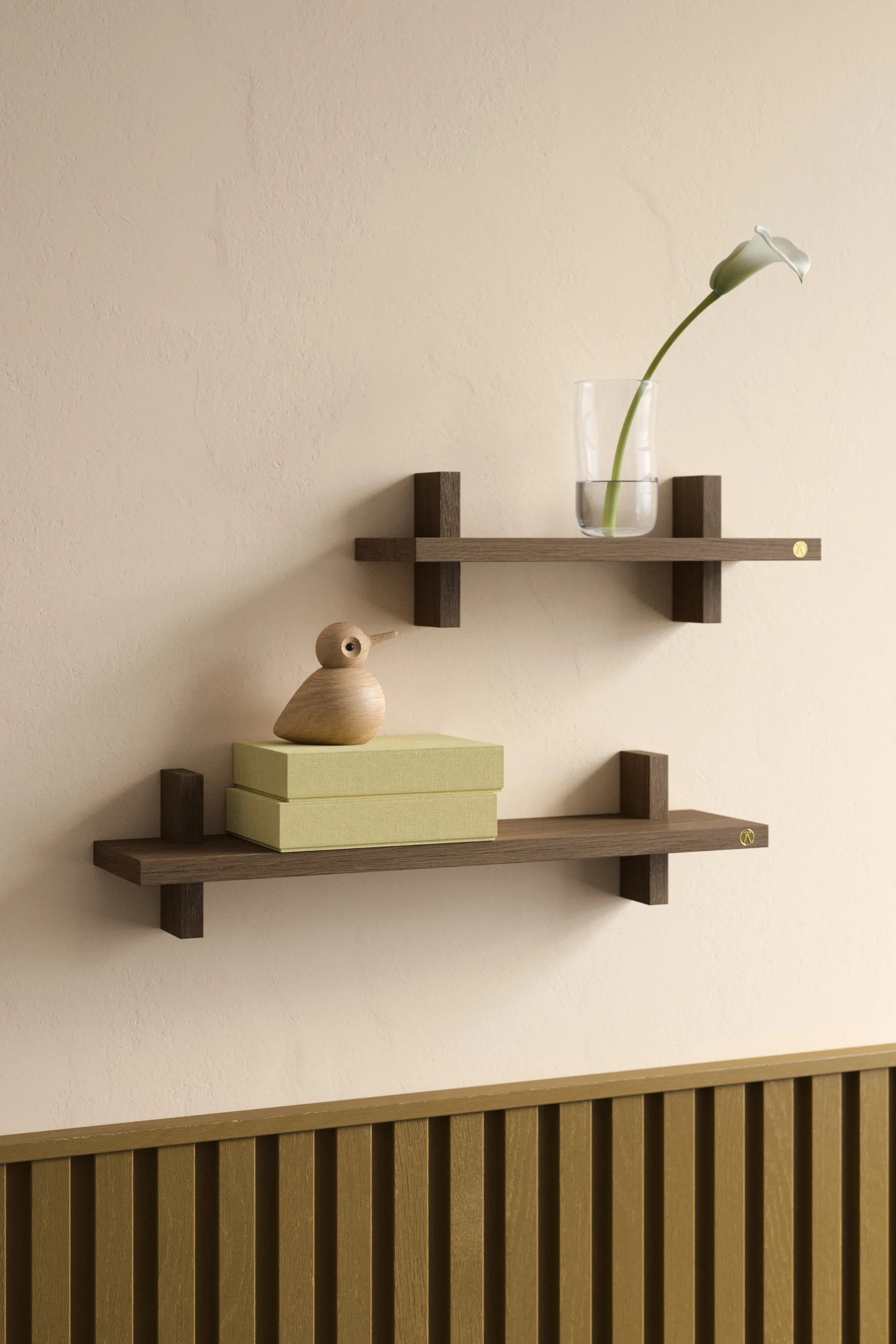 Dos estantes flotantes de madera oscura en una pared beige, uno con un jarrón y una azucena, el otro con cajas verdes y un pájaro de madera.