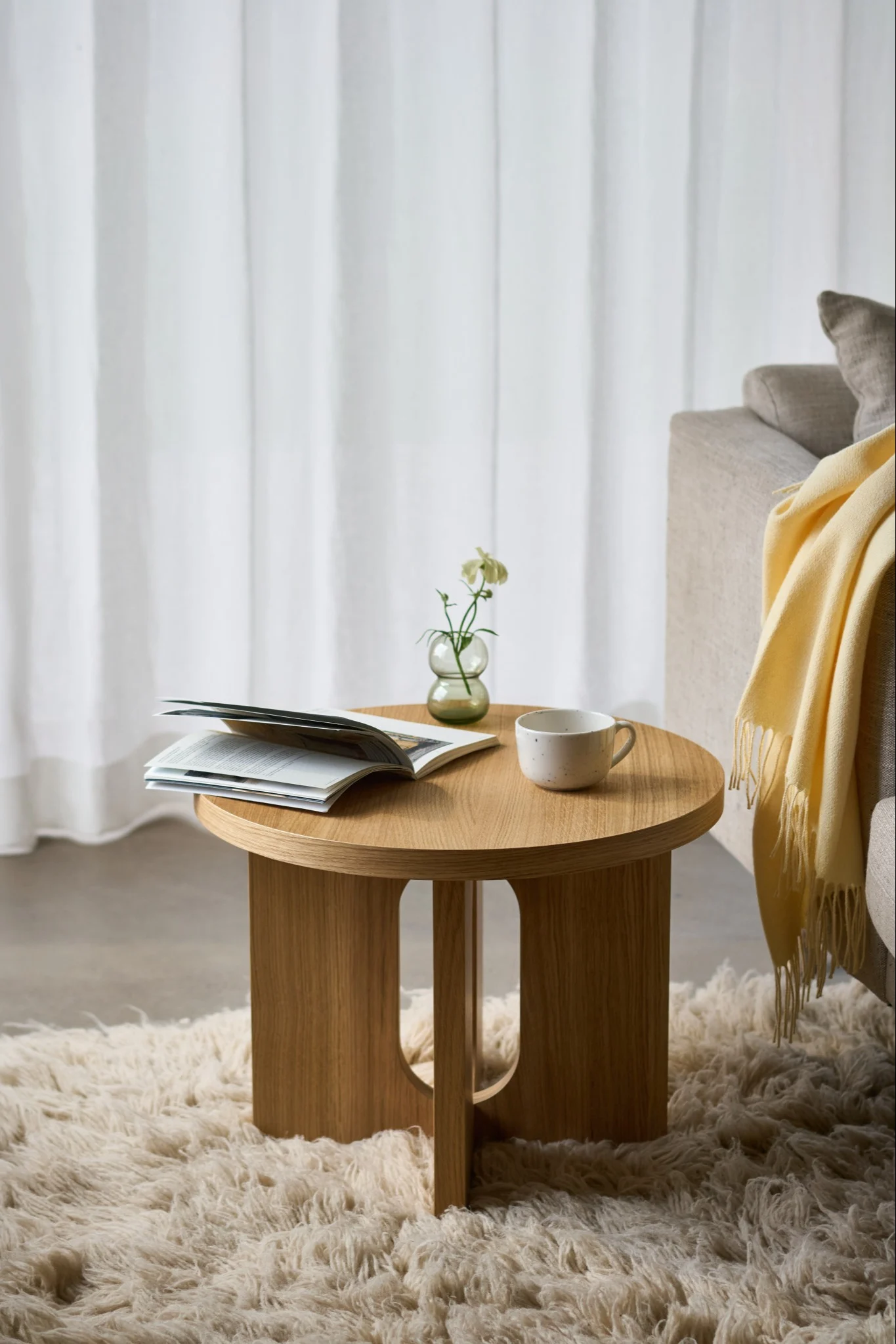 Mesa auxiliar redonda de madera con un libro abierto, una taza y un pequeño jarrón con una flor blanca sobre una alfombra de pelo largo, junto a un sofá.