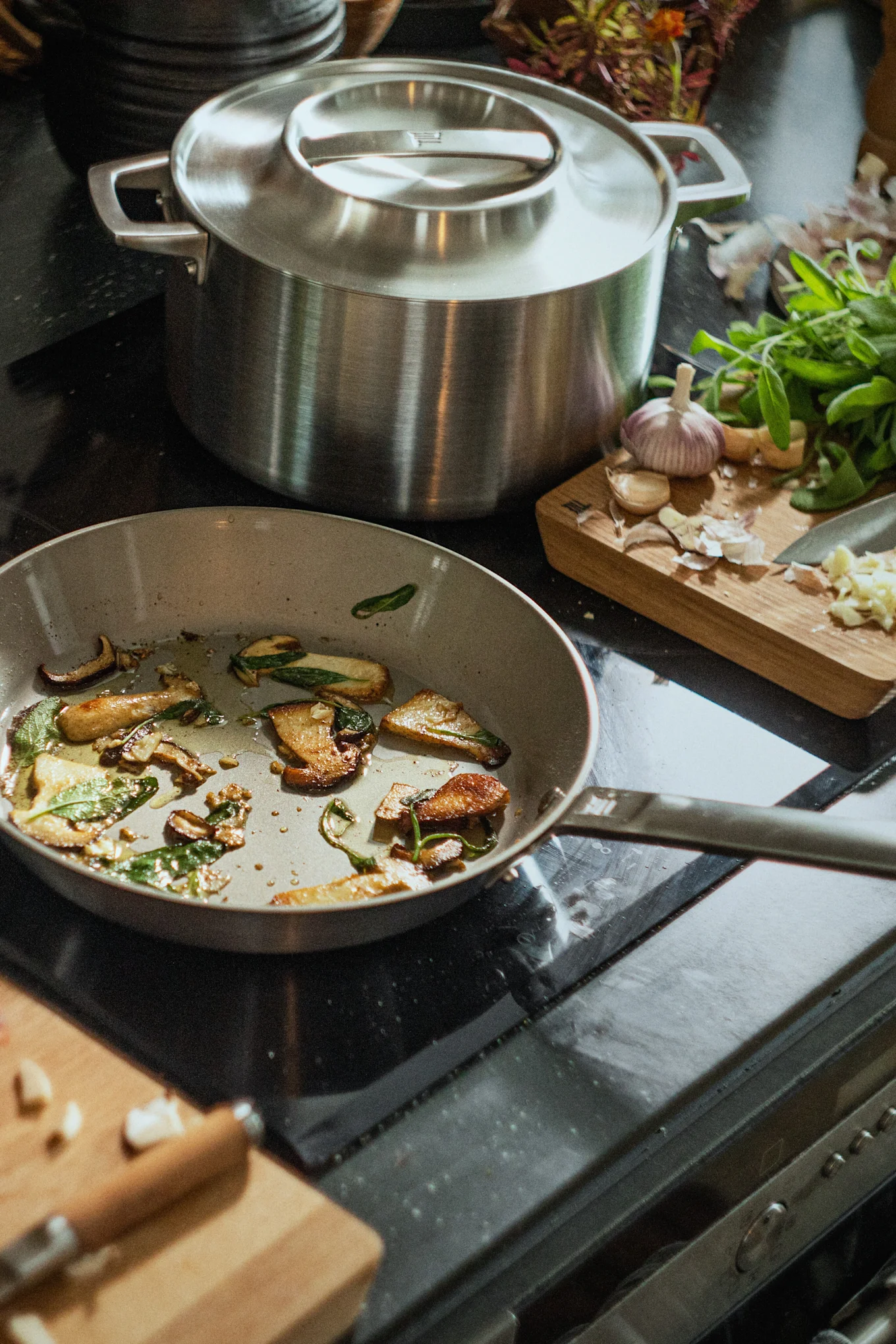 Una sartén con champiñones salteados y salvia en una estufa junto a una olla grande y una tabla de cortar con ajo.