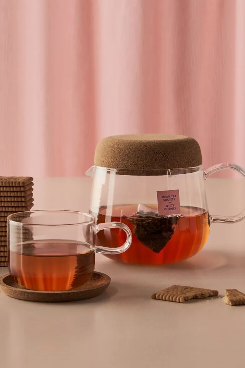Tetera de cristal con tapa de corcho y bolsita de té, una taza de té y galletas de chocolate sobre fondo rosa claro.