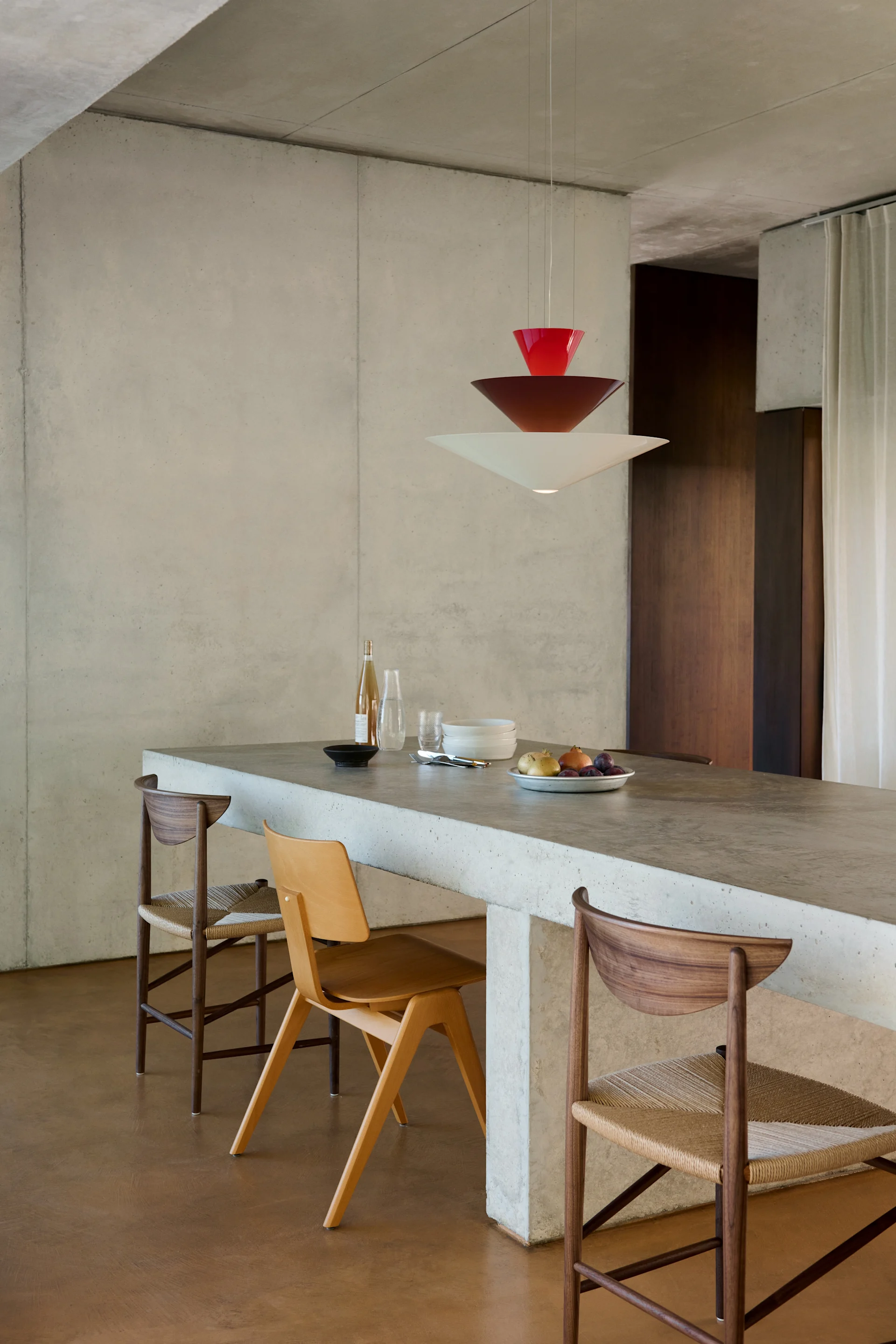 Comedor moderno con isla de hormigón, tres sillas de madera y una lámpara colgante escalonada roja y blanca.