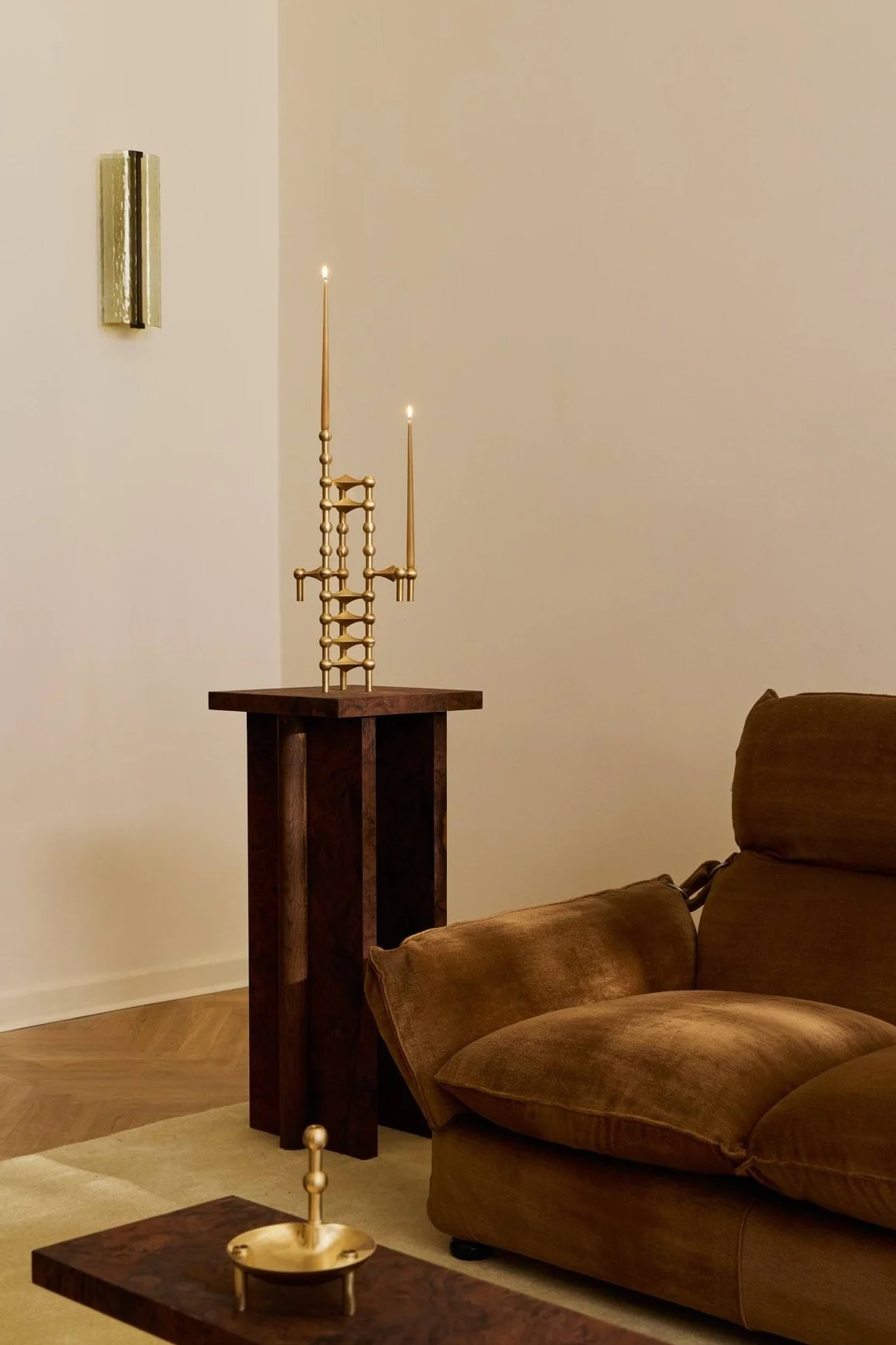 Un candelabro escultural de latón con dos velas encendidas sobre una mesa de pedestal de madera oscura, junto a un sofá de terciopelo marrón.