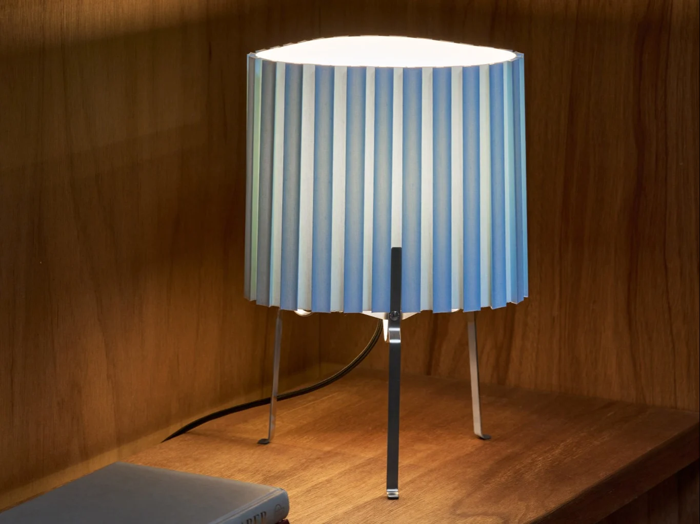 Lámpara de mesa con pantalla plisada de rayas azules y blancas sobre un estante de madera, iluminando el área.
