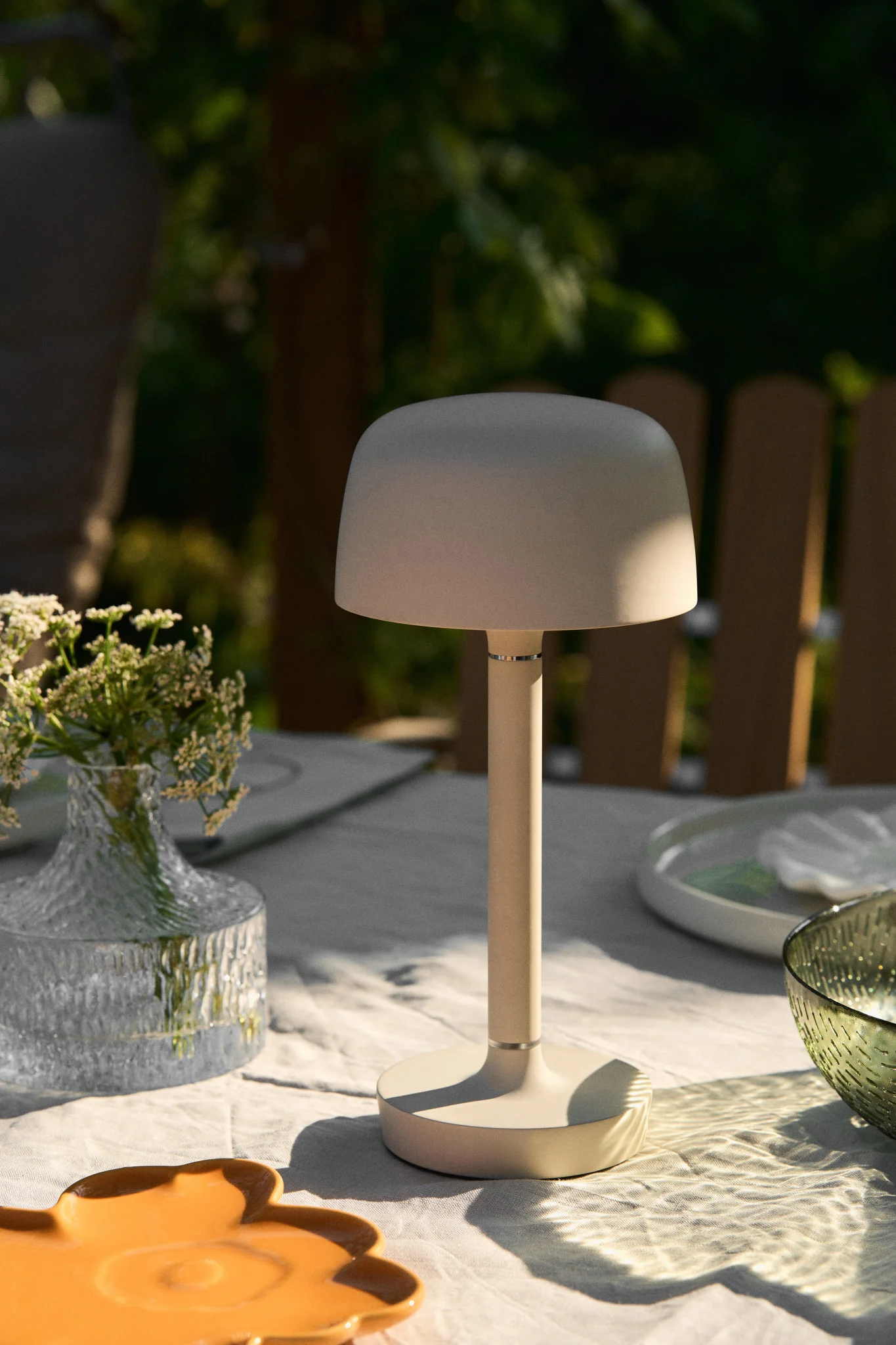 Una lámpara de mesa beige con forma de seta sobre una mesa de comedor blanca al aire libre con flores y platos.