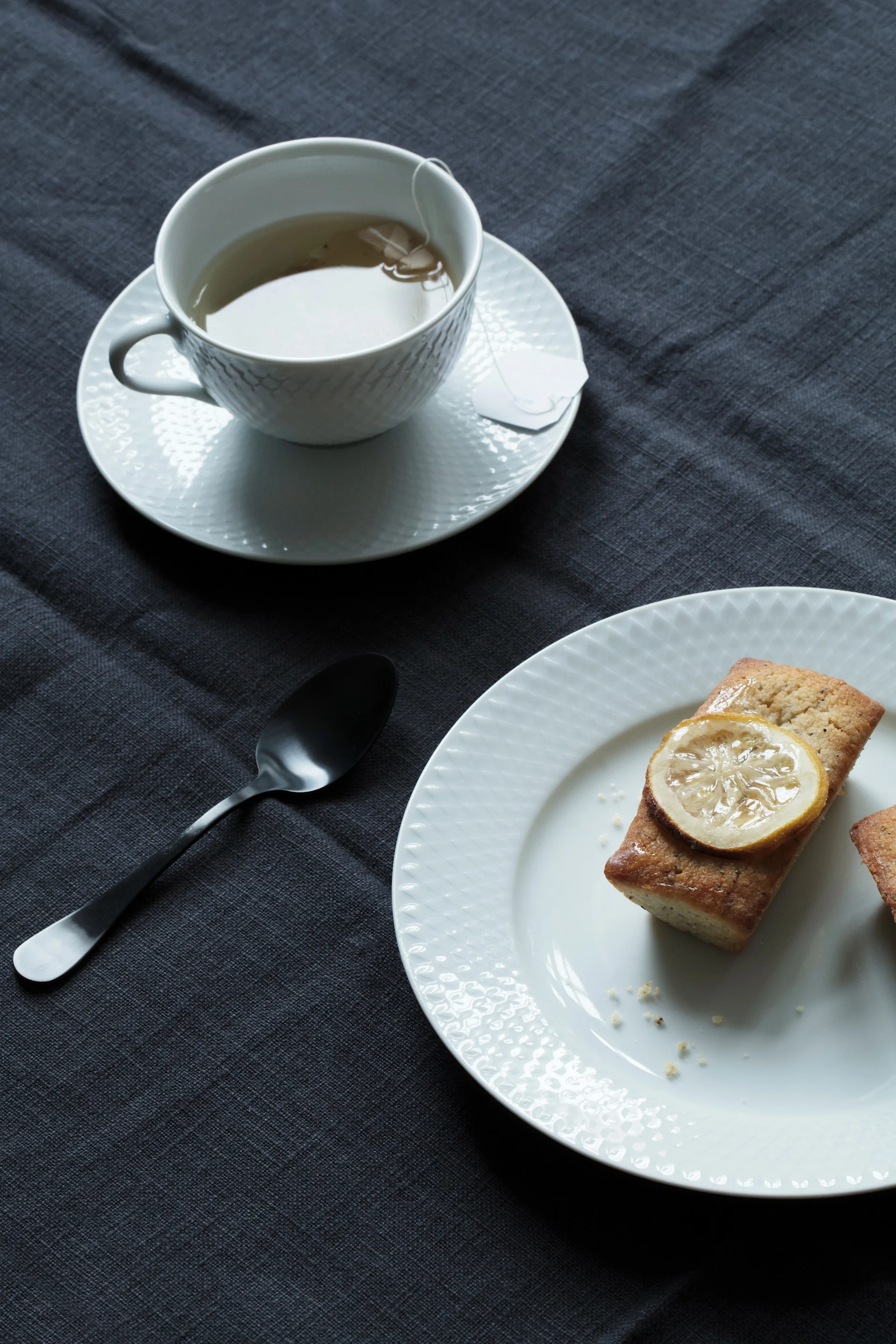 Una taza de té, una cuchara y dos pasteles de limón con limón confitado en un plato blanco, sobre un mantel gris oscuro.