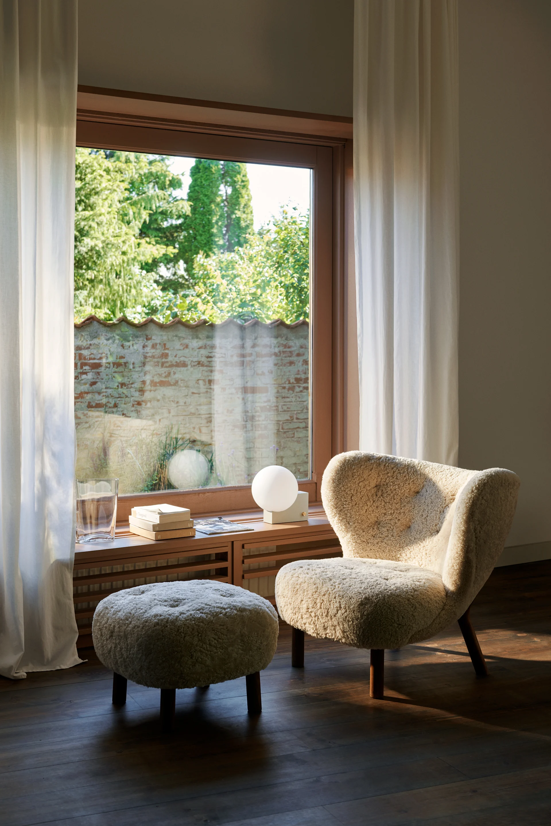 Sillón y otomana bouclé color crema junto a una ventana soleada. En el alféizar de madera: un vaso, libros y una lámpara esférica.