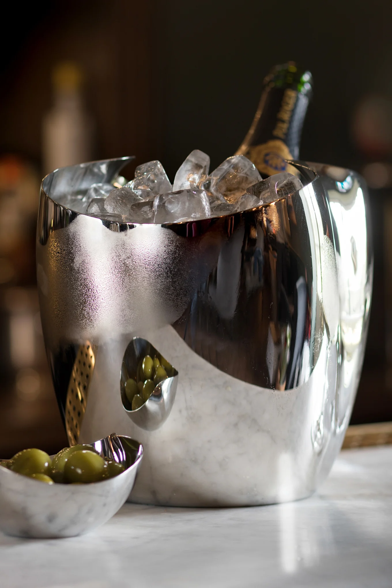Un cubo de hielo plateado reflectante con una botella de champán y hielo, junto a dos cuencos pequeños con aceitunas verdes sobre mármol.