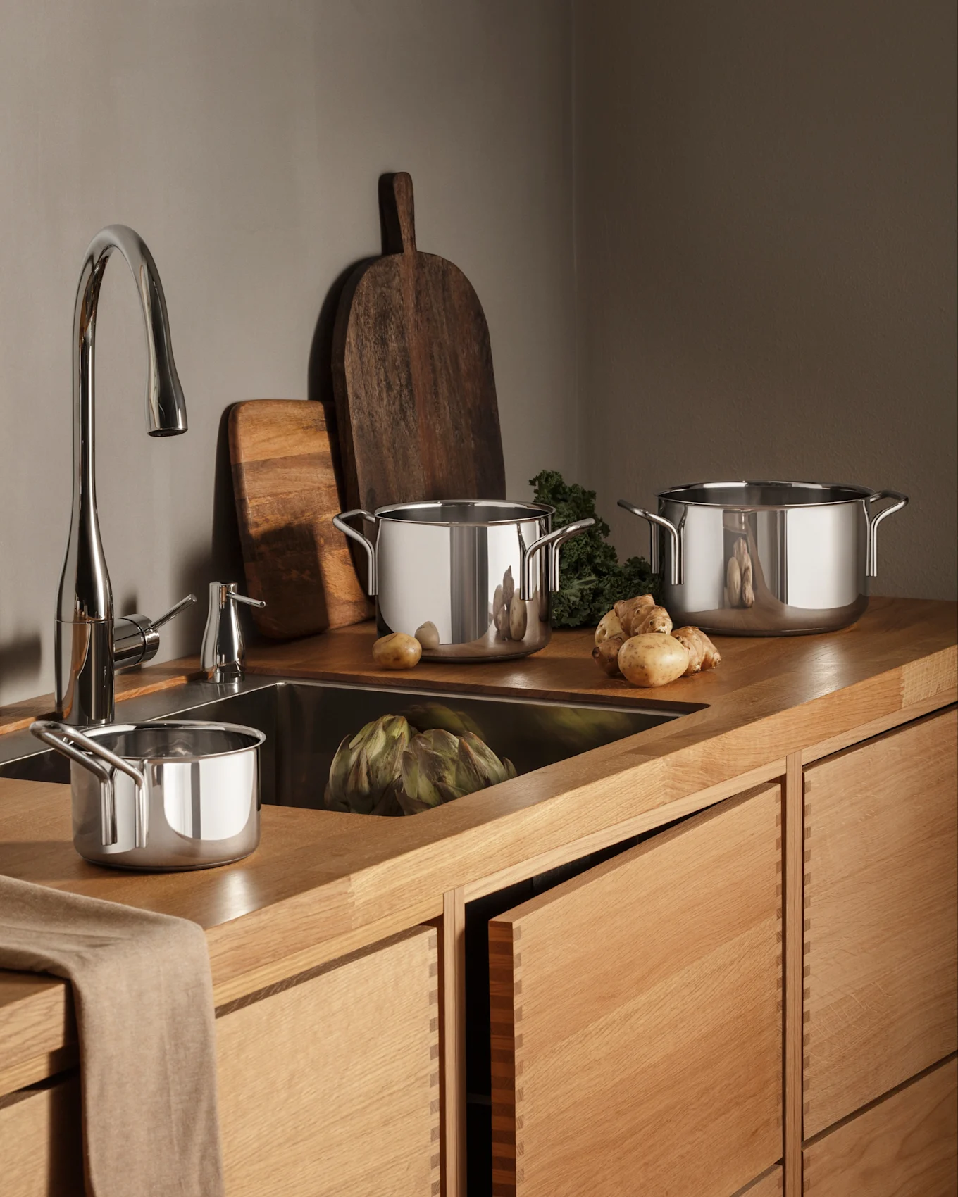 Una cocina moderna con encimeras y gabinetes de madera, un grifo cromado y ollas de acero inoxidable, productos frescos incluyendo alcachofa y patatas.
