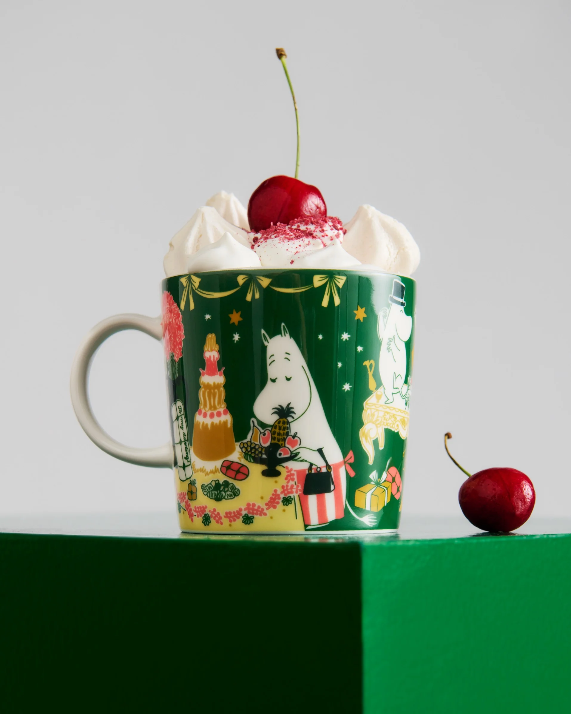 Taza verde oscuro con personajes Moomin, con nata montada, chispas rojas y una cereza. Otra cereza está sobre la superficie verde.