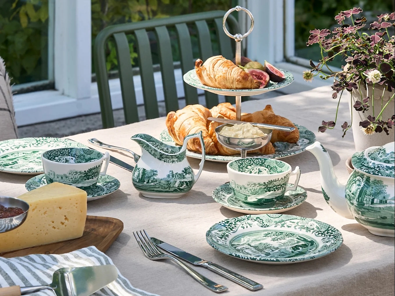 Una mesa exterior puesta con vajilla verde y blanca, cruasanes, queso y una tetera, en un jardín.