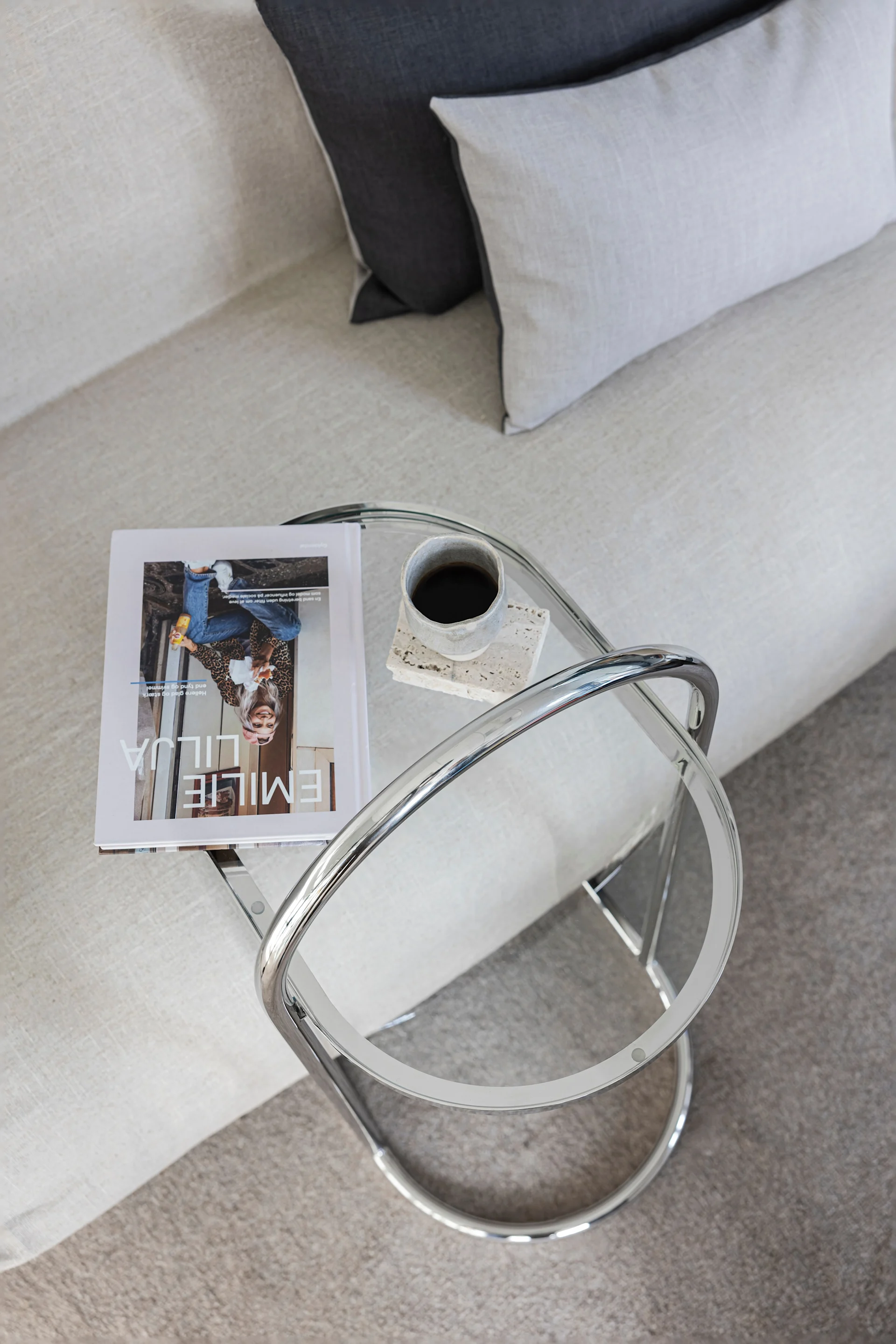 Vista aérea de una sala de estar moderna con un sofá claro, cojines y una mesa auxiliar de vidrio y cromo con una revista y café.
