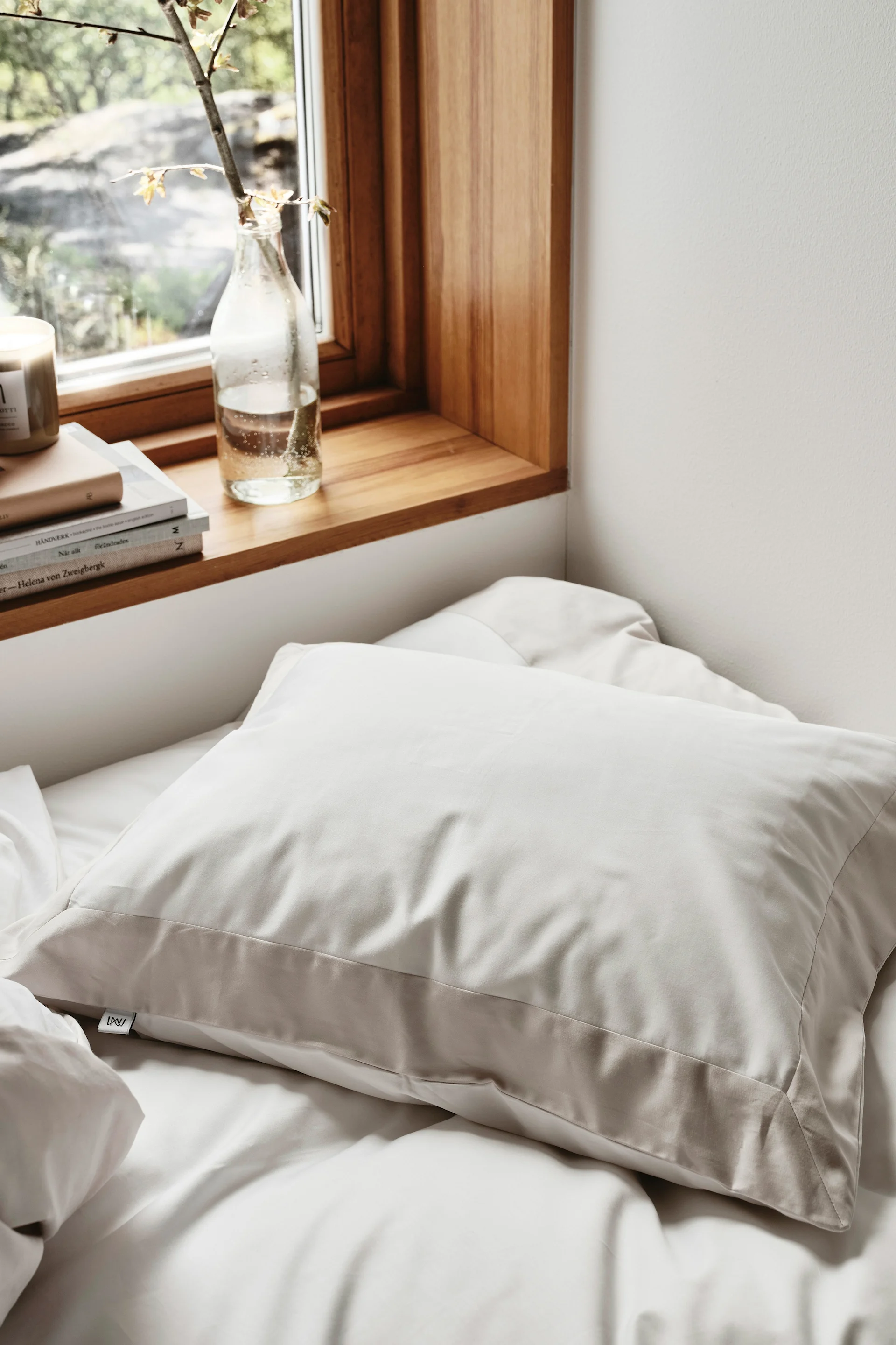 Una almohada beige claro en una cama blanca, junto a una ventana con alféizar de madera, libros y una rama de planta en una botella.