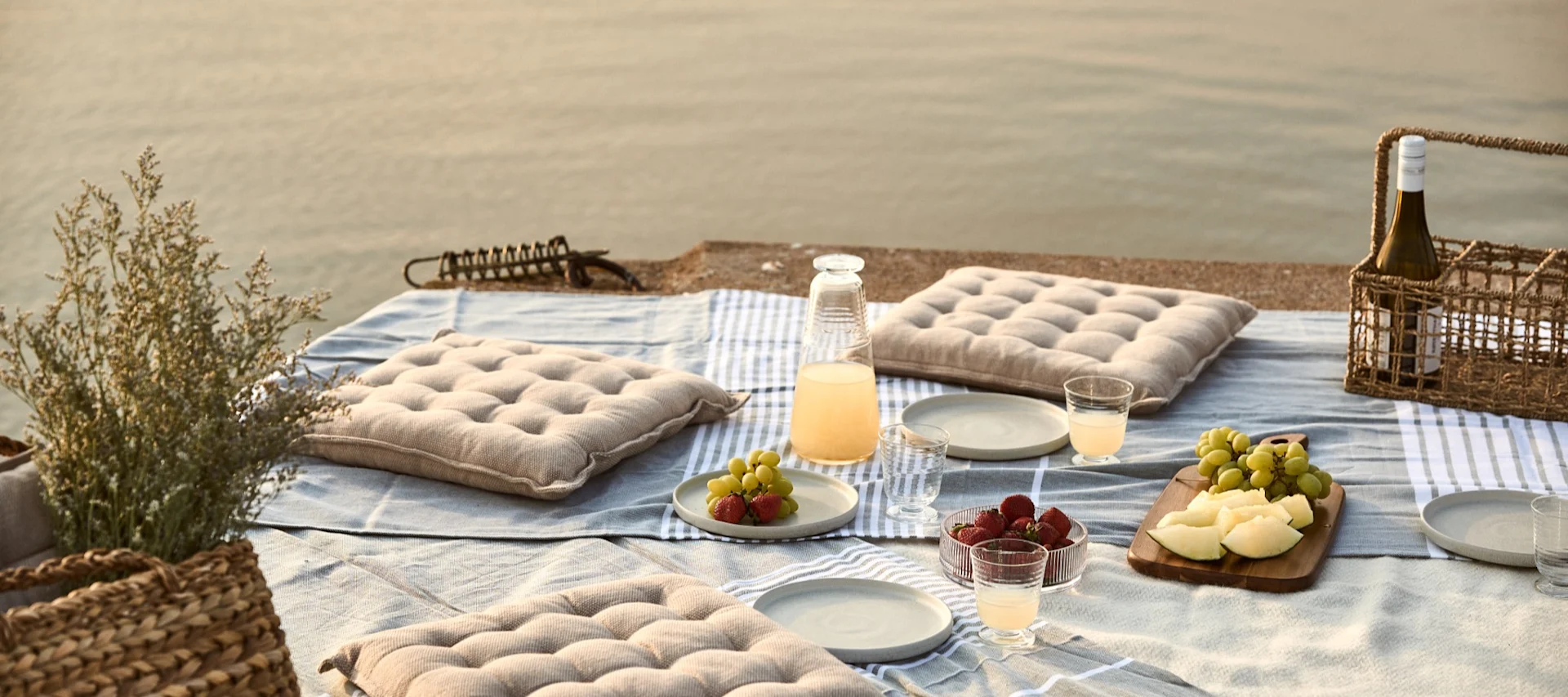 Picnic junto al agua con cojines, fruta y bebidas sobre una manta.