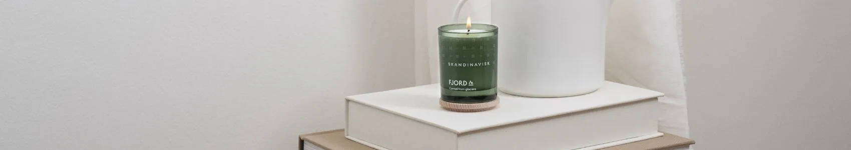 Un libro titulado 'NOTES FROM THE FELLS' sobre una manta gris junto a dos velas de estilo escandinavo, una negra encendida y otra marrón con tapa de madera.
