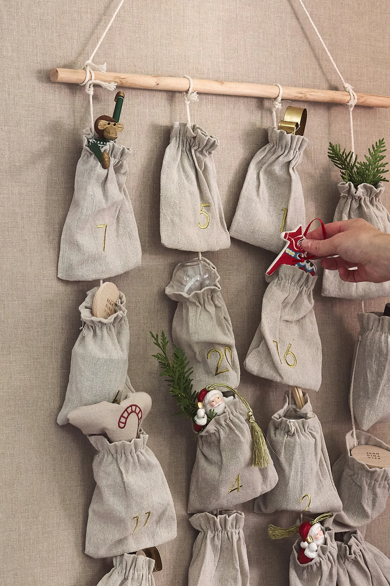 Calendario de Adviento de tela con bolsas numeradas colgando de una barra de madera; una mano coloca un caballo Dala rojo.