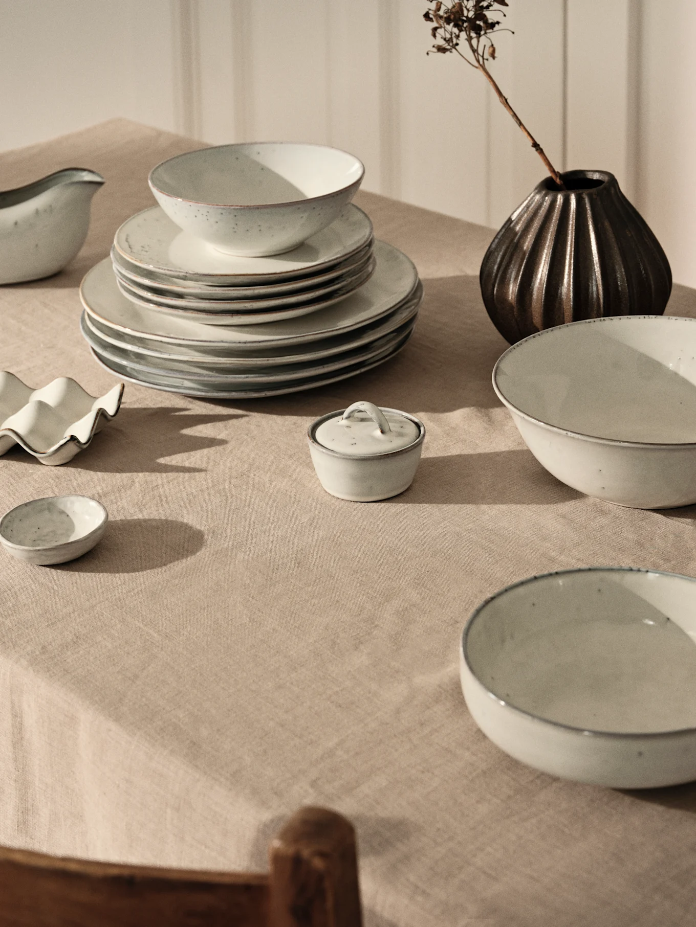Vajilla de cerámica gris claro con platos y cuencos apilados en mantel de lino beige, junto a jarrón oscuro con rama seca.
