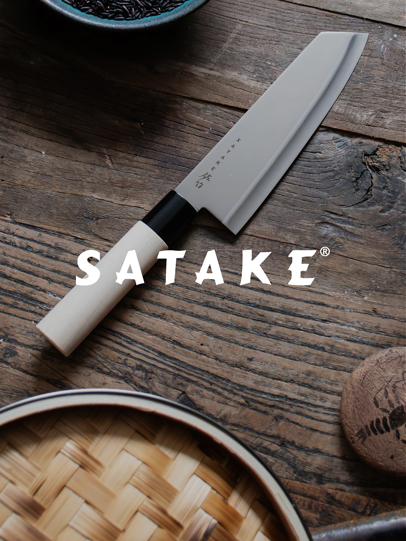 Un cuchillo de chef Satake con mango claro y hoja gris sobre una mesa de madera, junto a arroz negro y una vaporera de bambú.